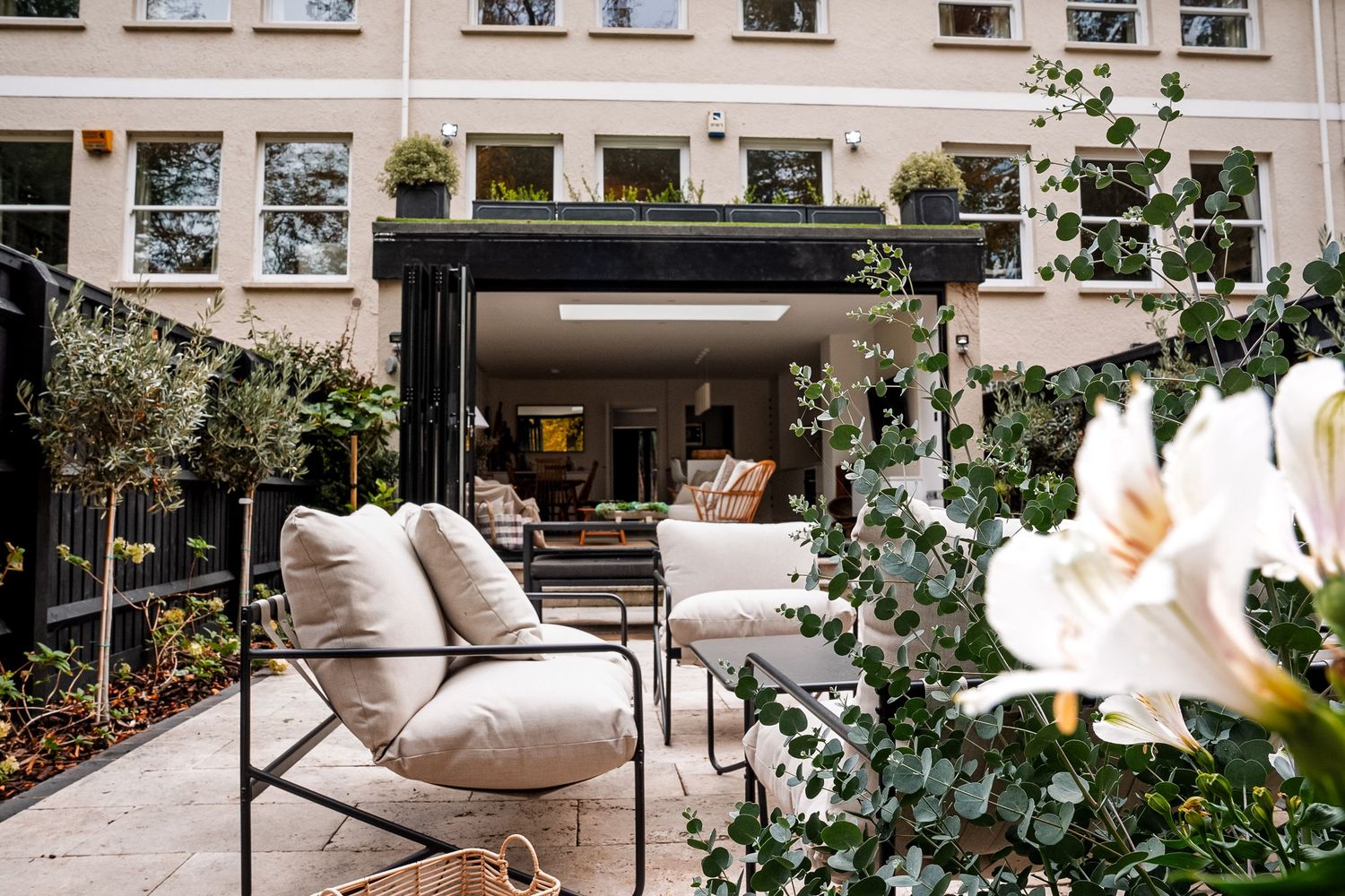 Elegant outdoor patio with white cushioned chairs, black metal frames, lush greenery, and open glass doors leading to a bright interior.