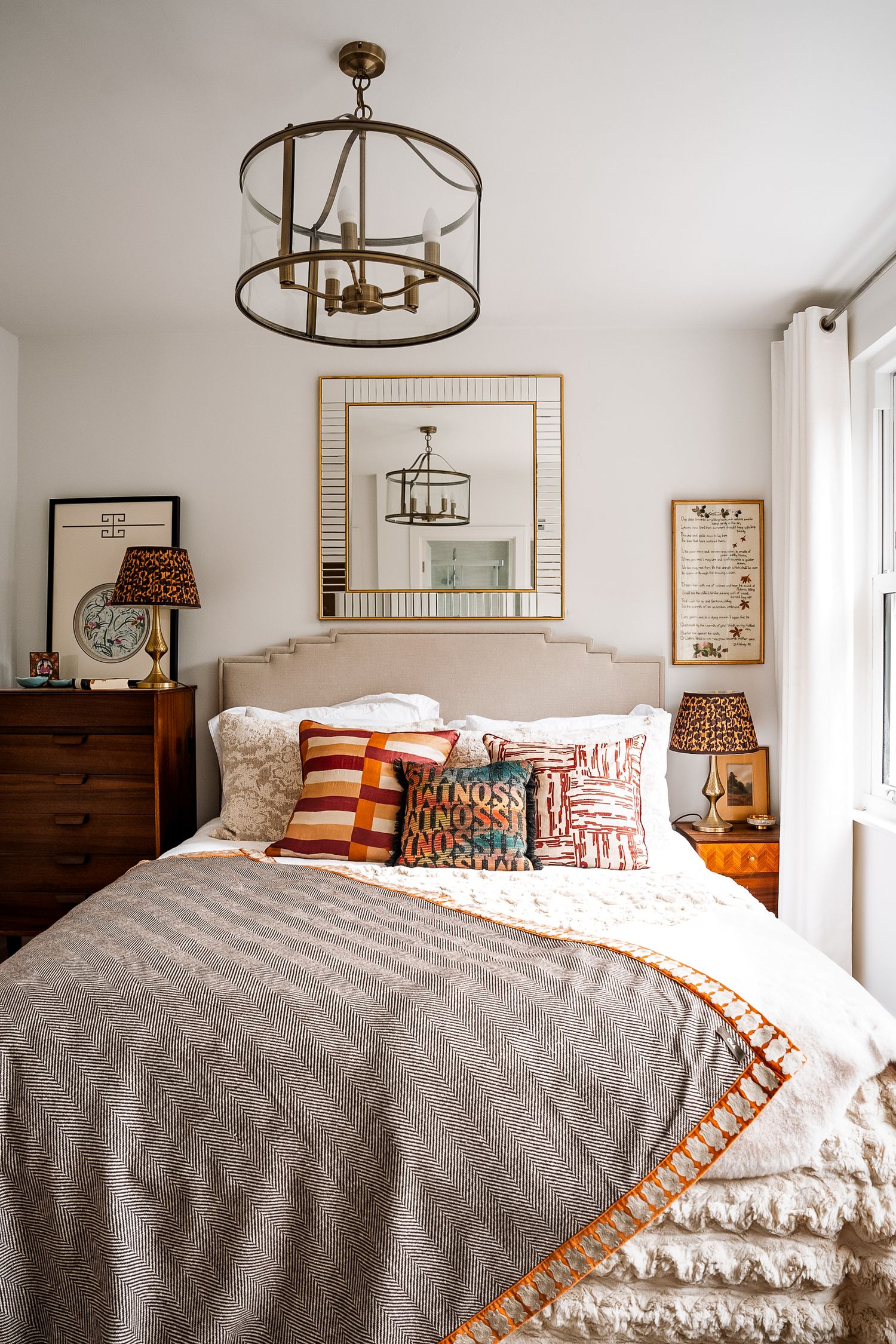 Cozy bedroom with herringbone throw, colorful pillows, upholstered headboard, wood nightstands, and brass lantern chandelier.