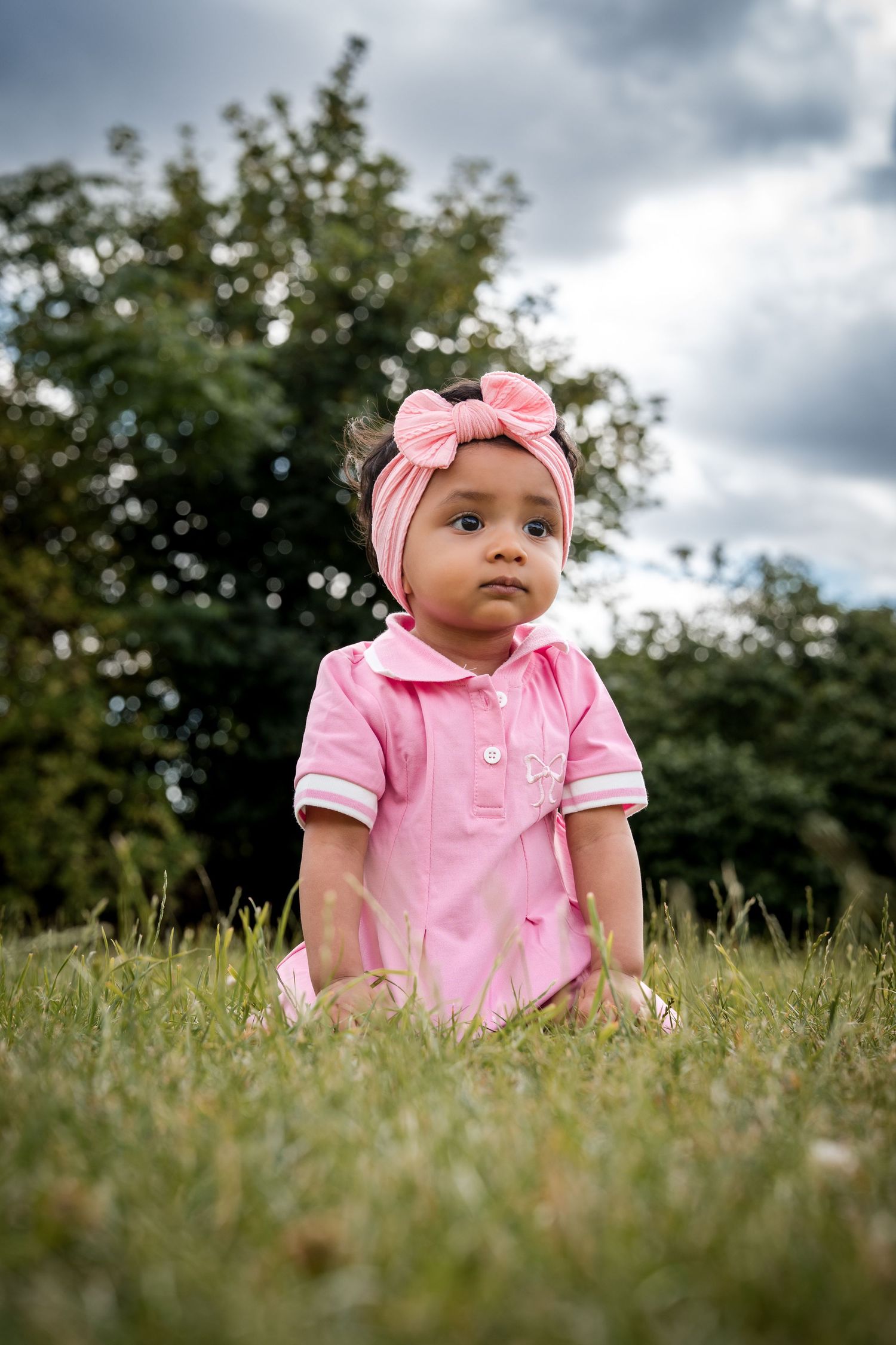 Adorable baby girl in pink dress and bow headband sitting on green grass outdoors with trees in background.