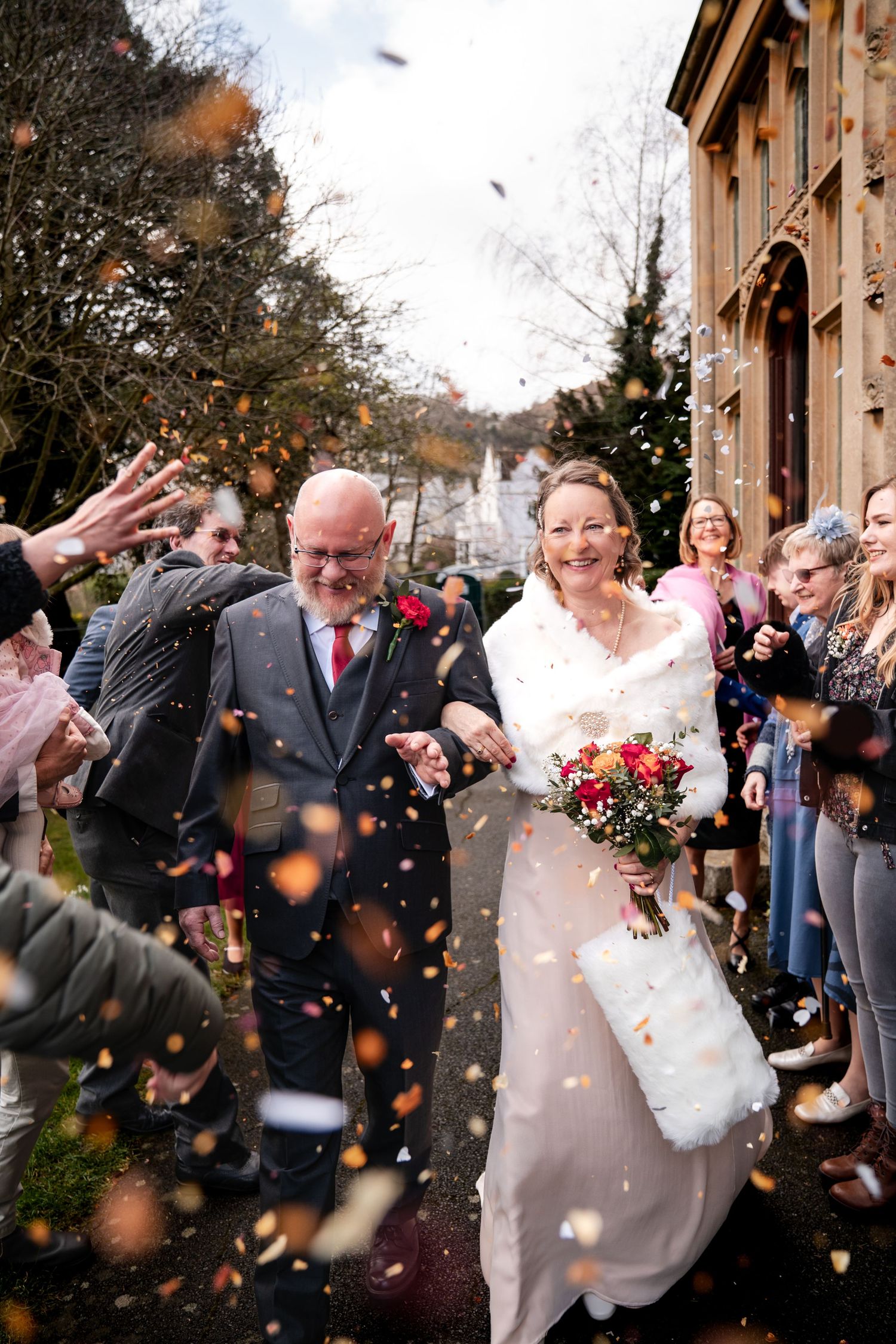 Bride and groom walk through confetti shower surrounded by smiling guests outside a historic building on their wedding day.