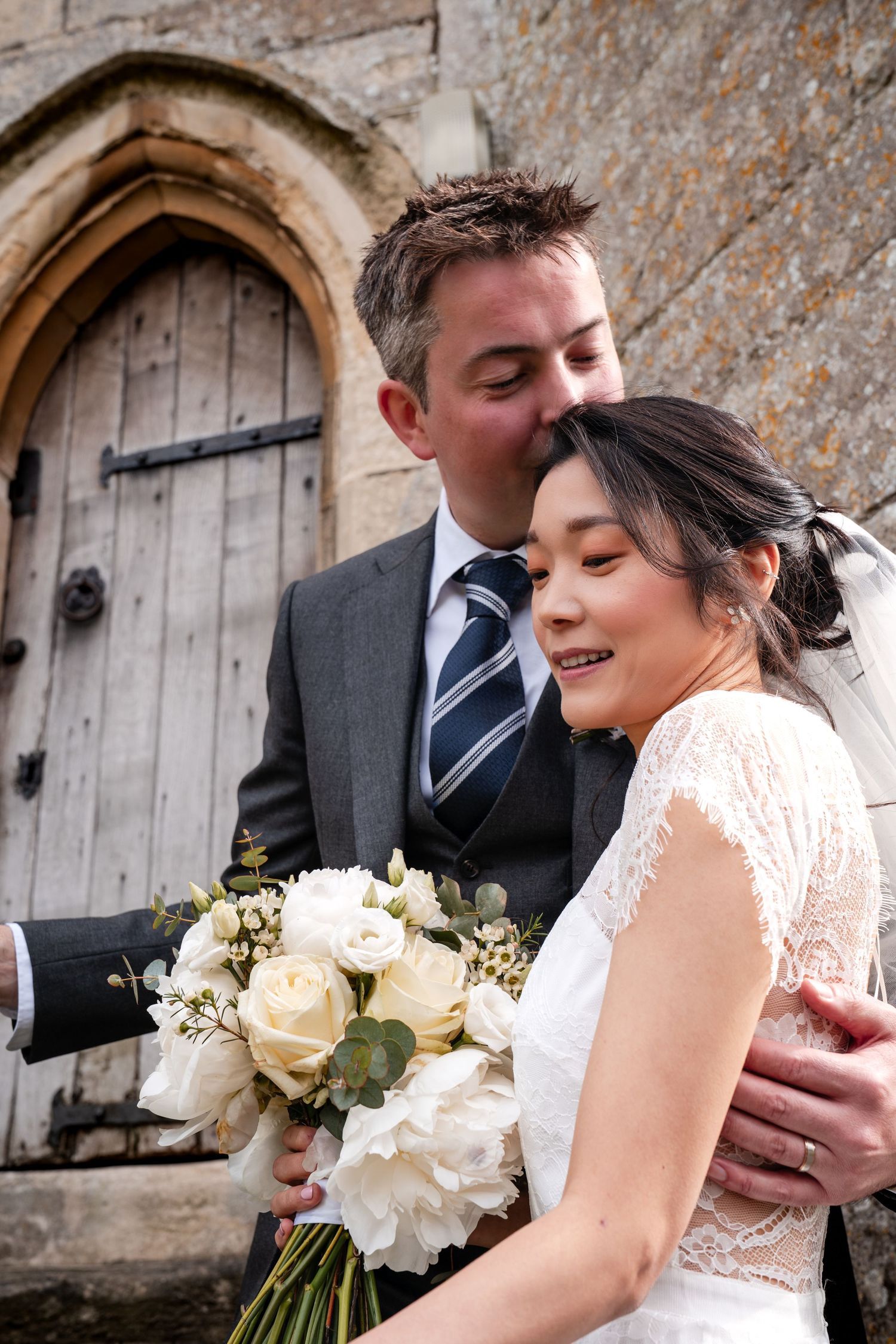Groom kissing bride on head outside stone church, bride holding white rose bouquet in lace wedding dress.