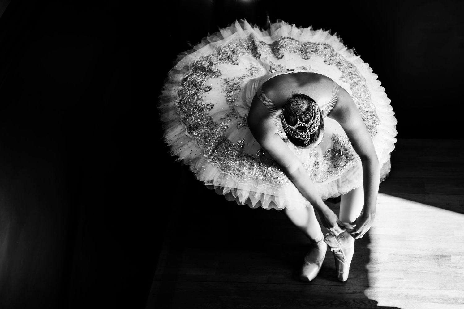 Black and white photo of a ballerina in a tutu bending gracefully to adjust her pointe shoes on a dark stage.