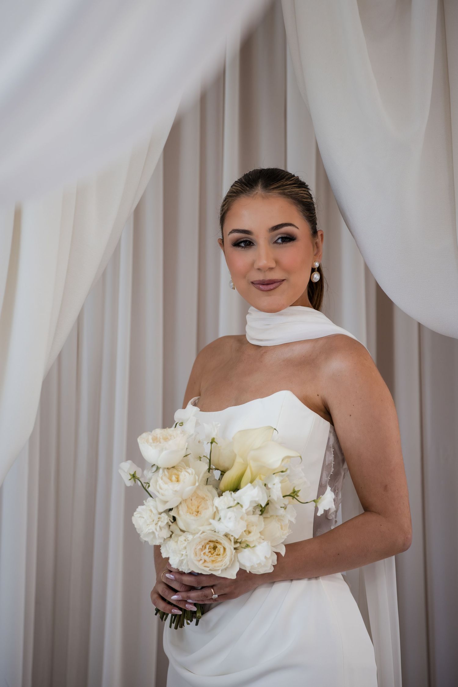 Elegant bride in strapless white gown holding bouquet of white roses and calla lilies, posing before draped curtains.