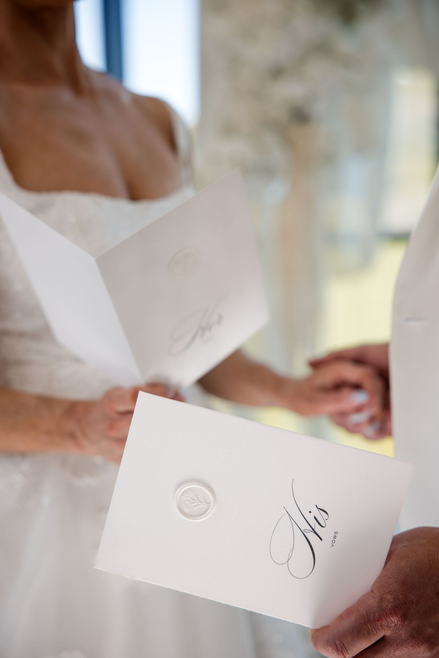 Bride and groom holding white wedding vow booklets with wax seals, labeled His and Hers, during ceremony.