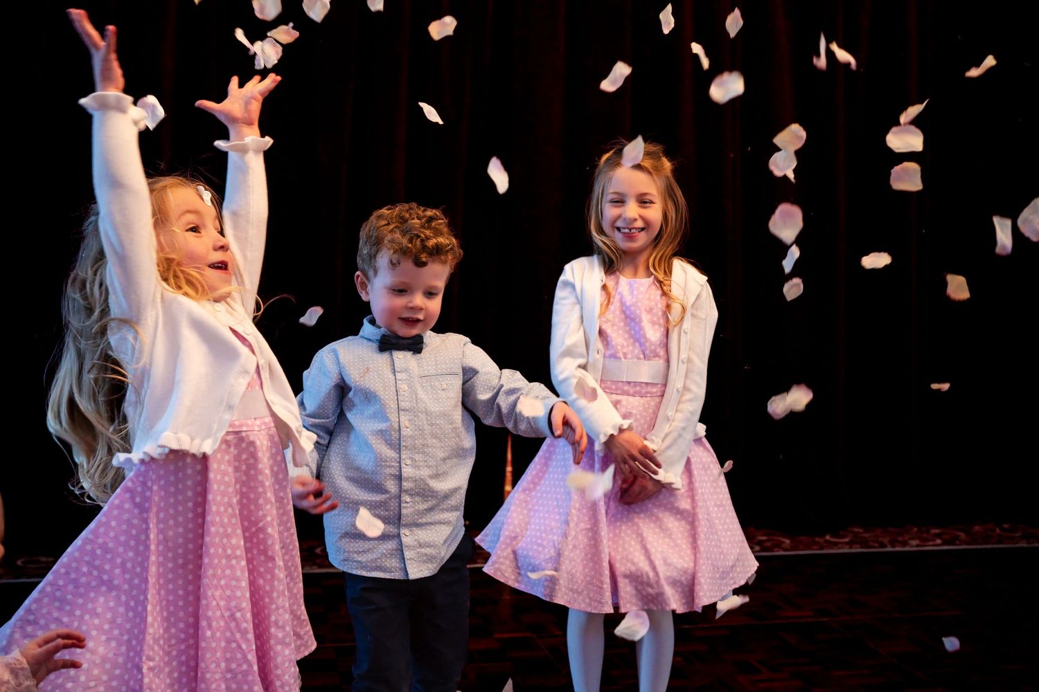 Three young children in formal attire celebrate joyfully as confetti falls around them on a stage.