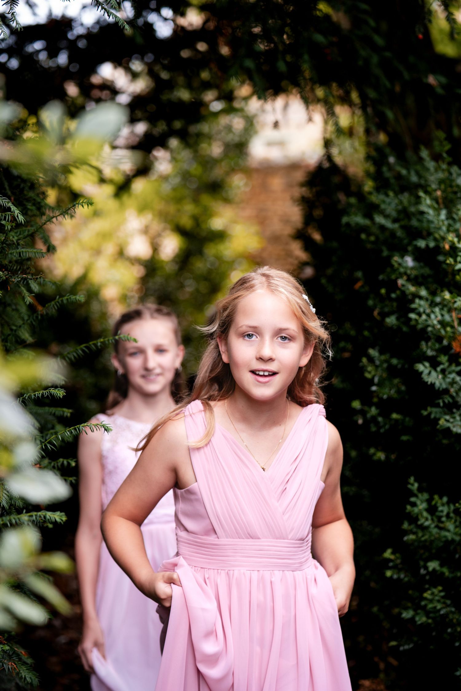 Two young girls in pink dresses walking through a lush green garden archway, smiling and looking elegant.