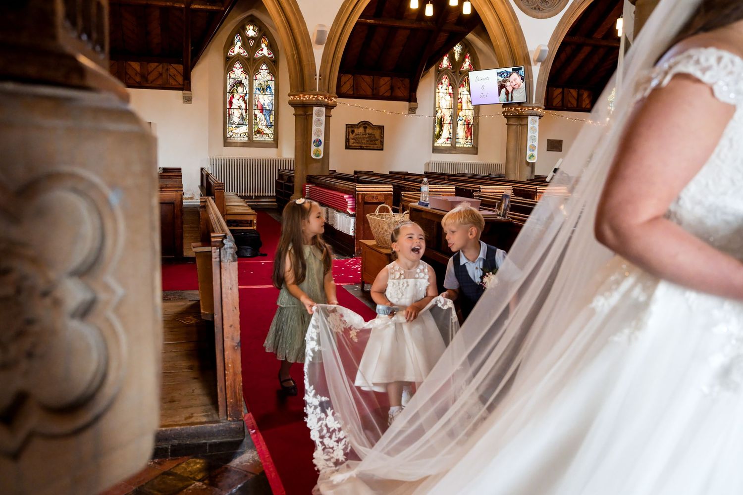 Children in formal attire admire a bride's wedding gown inside a traditional church with stained glass windows.