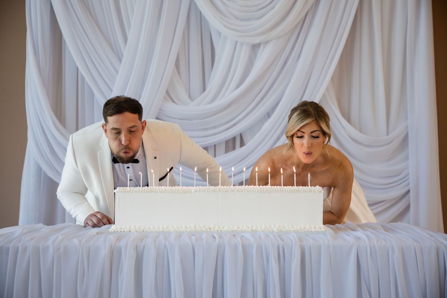 Bride and groom blowing out candles on white wedding cake at elegant reception with draped white backdrop.