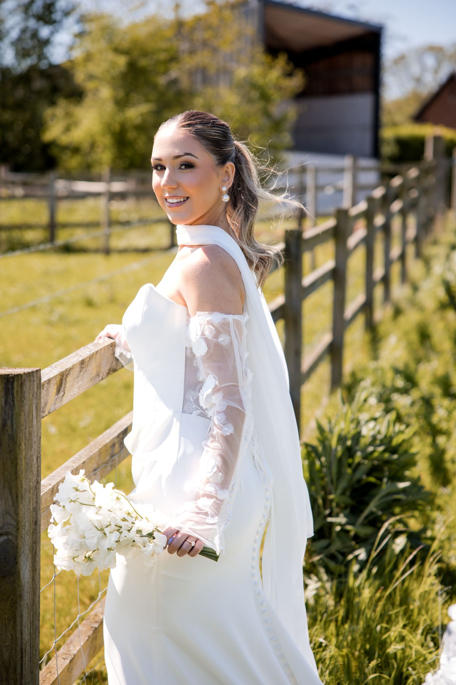 Smiling bride in white gown holding bouquet of white flowers by wooden fence on sunny day.