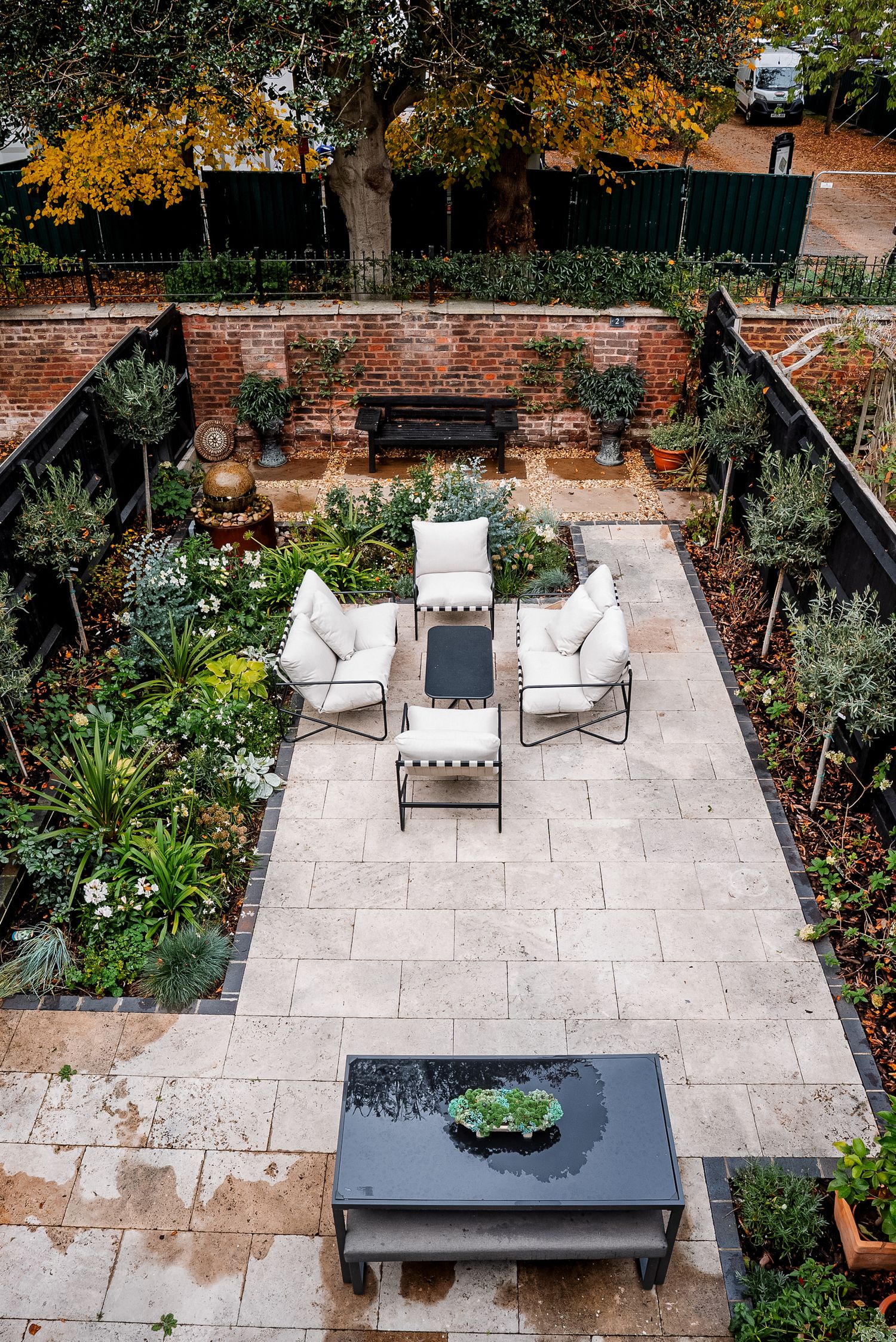 Aerial view of a elegant walled garden patio with white outdoor furniture, stone tiles, lush greenery, and brick walls.