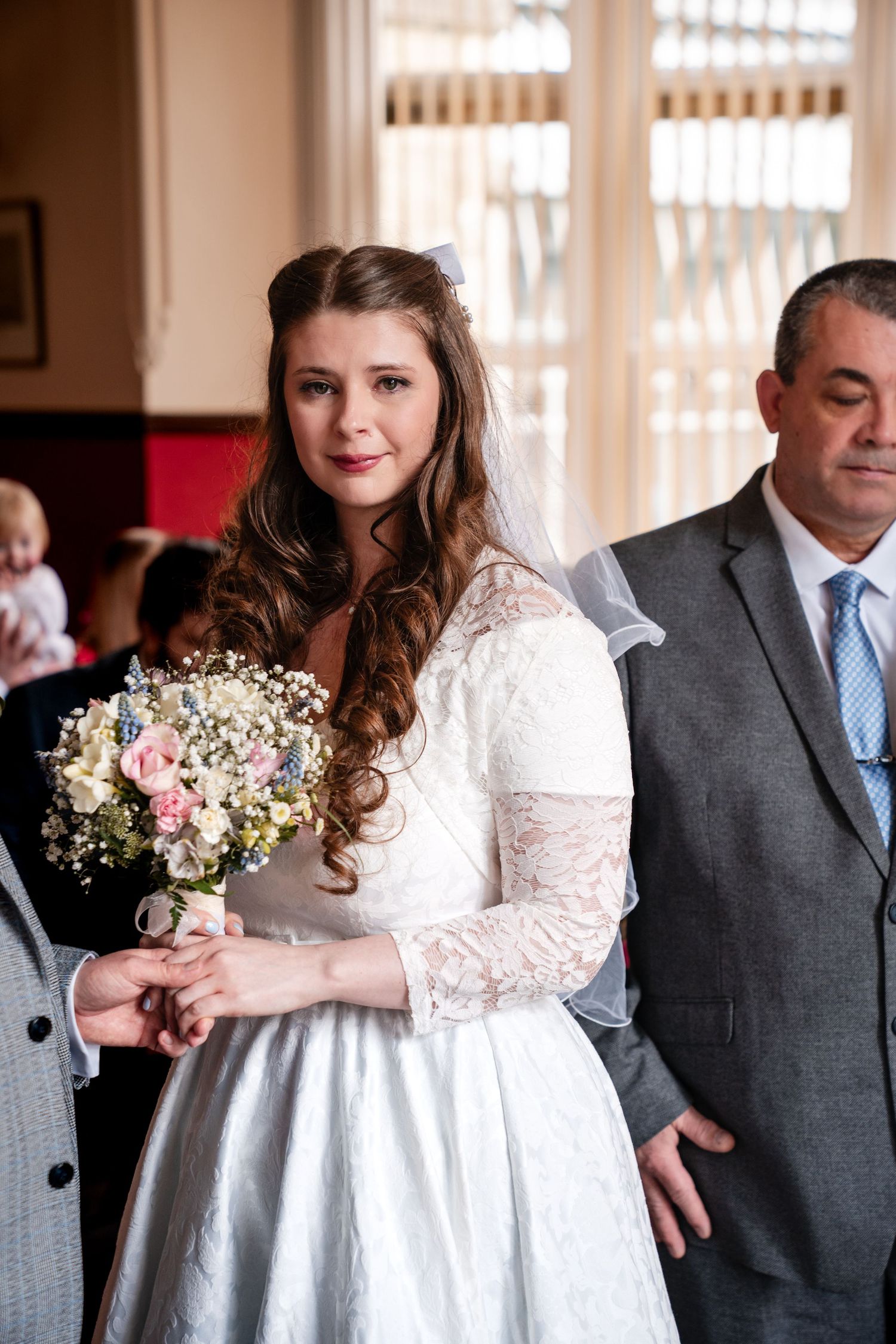 Bride in white lace dress holding floral bouquet walks down aisle accompanied by man in gray suit.