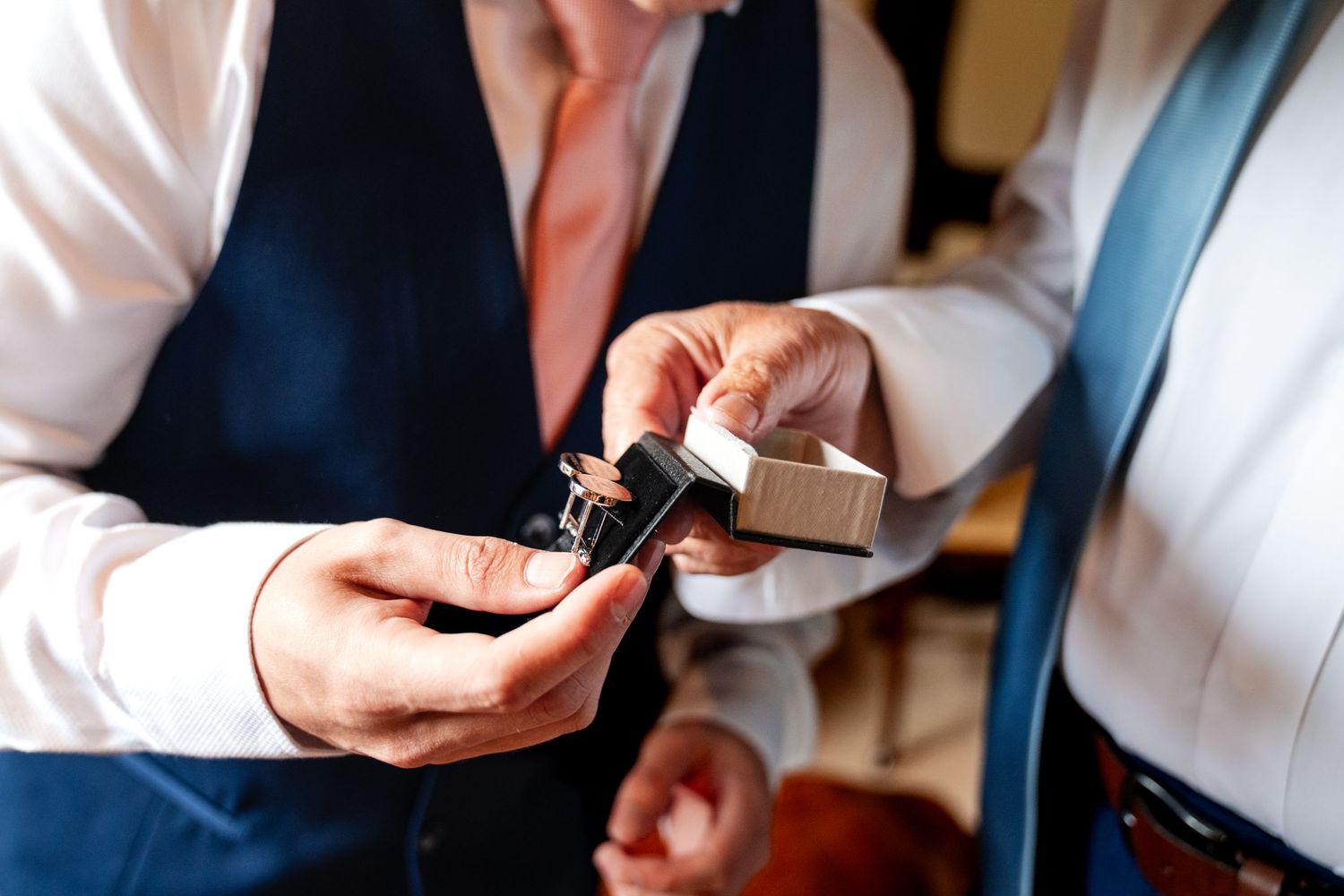 Two men examining cufflinks in a jewelry box, dressed in formal wedding attire with ties and vests.