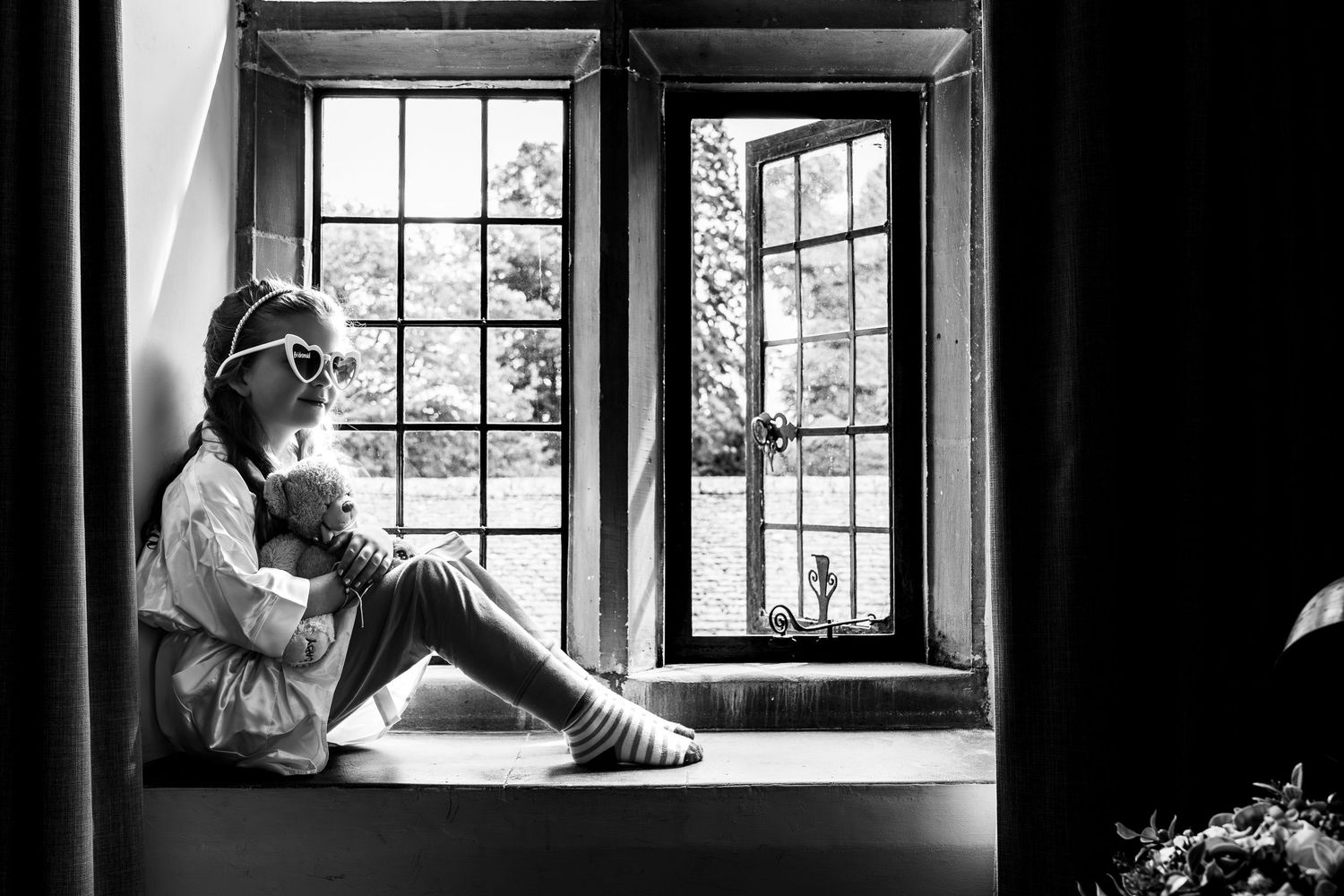 Black and white photo of a young girl sitting on a windowsill, gazing outside through vintage French windows.