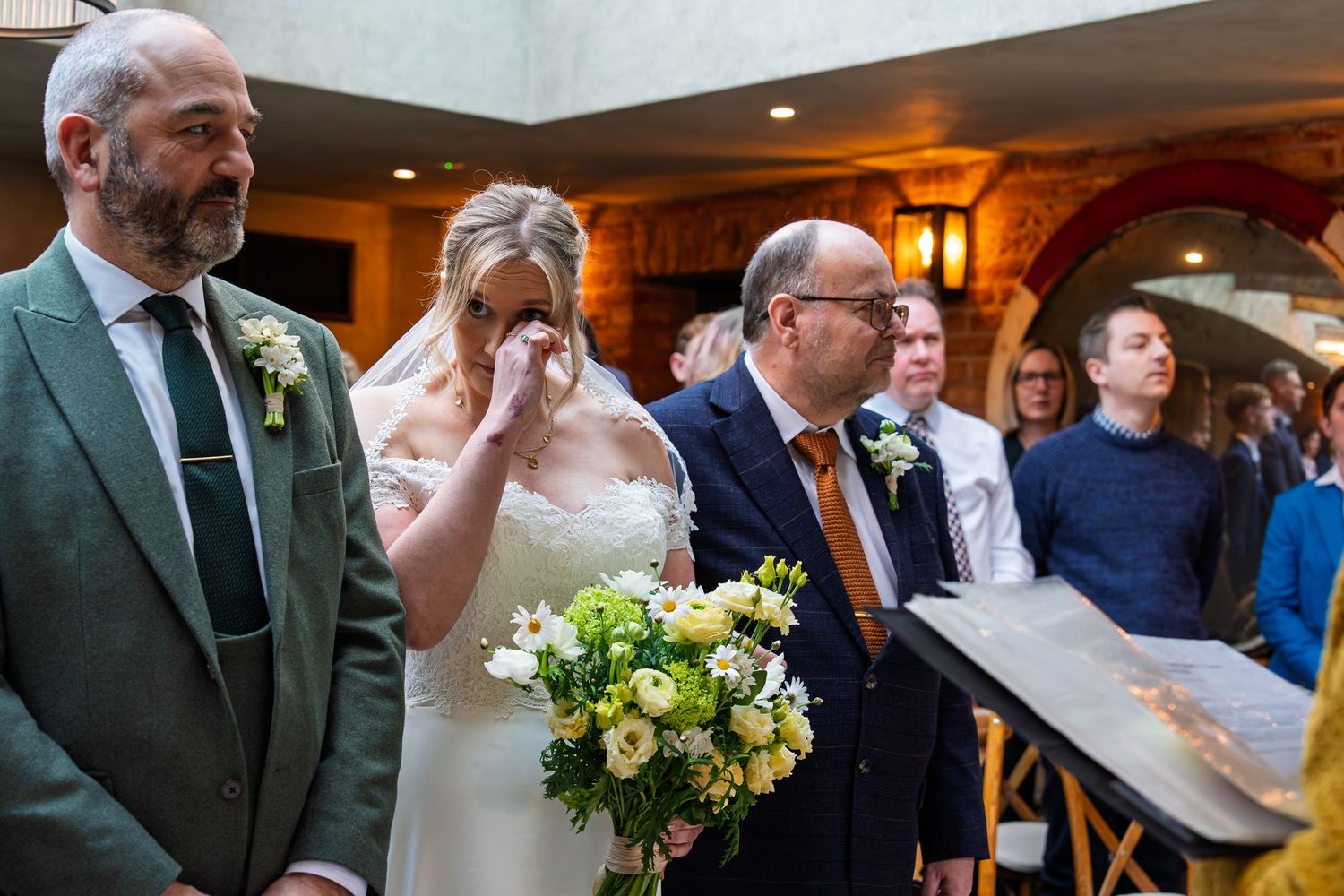 Emotional bride wiping tears holding white bouquet at wedding ceremony with guests and groom in suit beside her.