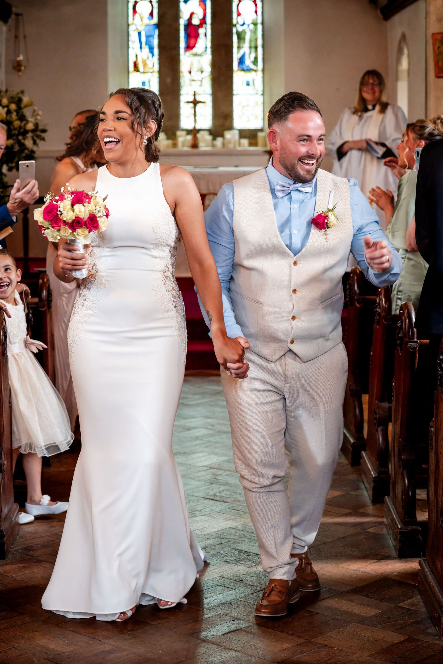 Joyful newlyweds walk down church aisle, bride in white gown holding bouquet, groom in grey vest, after wedding ceremony.