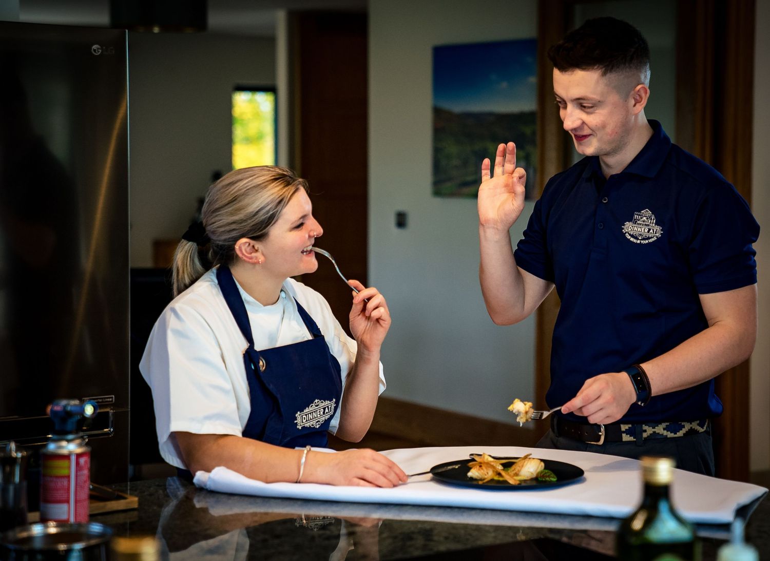 Two chefs tasting and discussing a gourmet dish plated on a black plate in a modern kitchen.