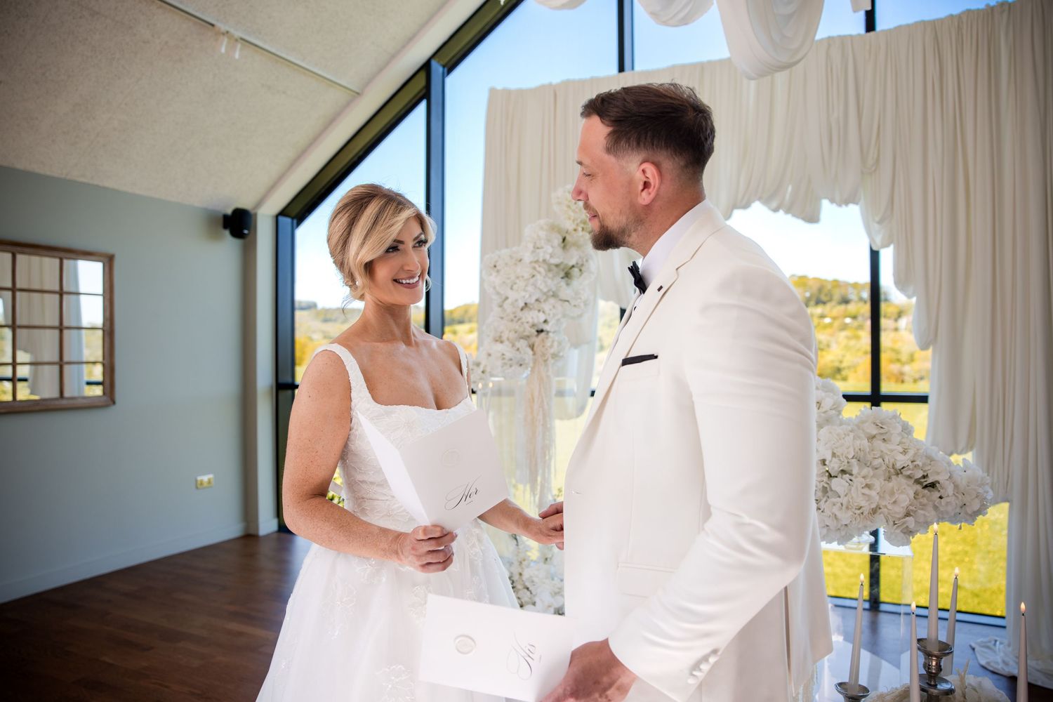 Bride and groom exchange vows at elegant white wedding ceremony venue with floral arrangements and large windows.