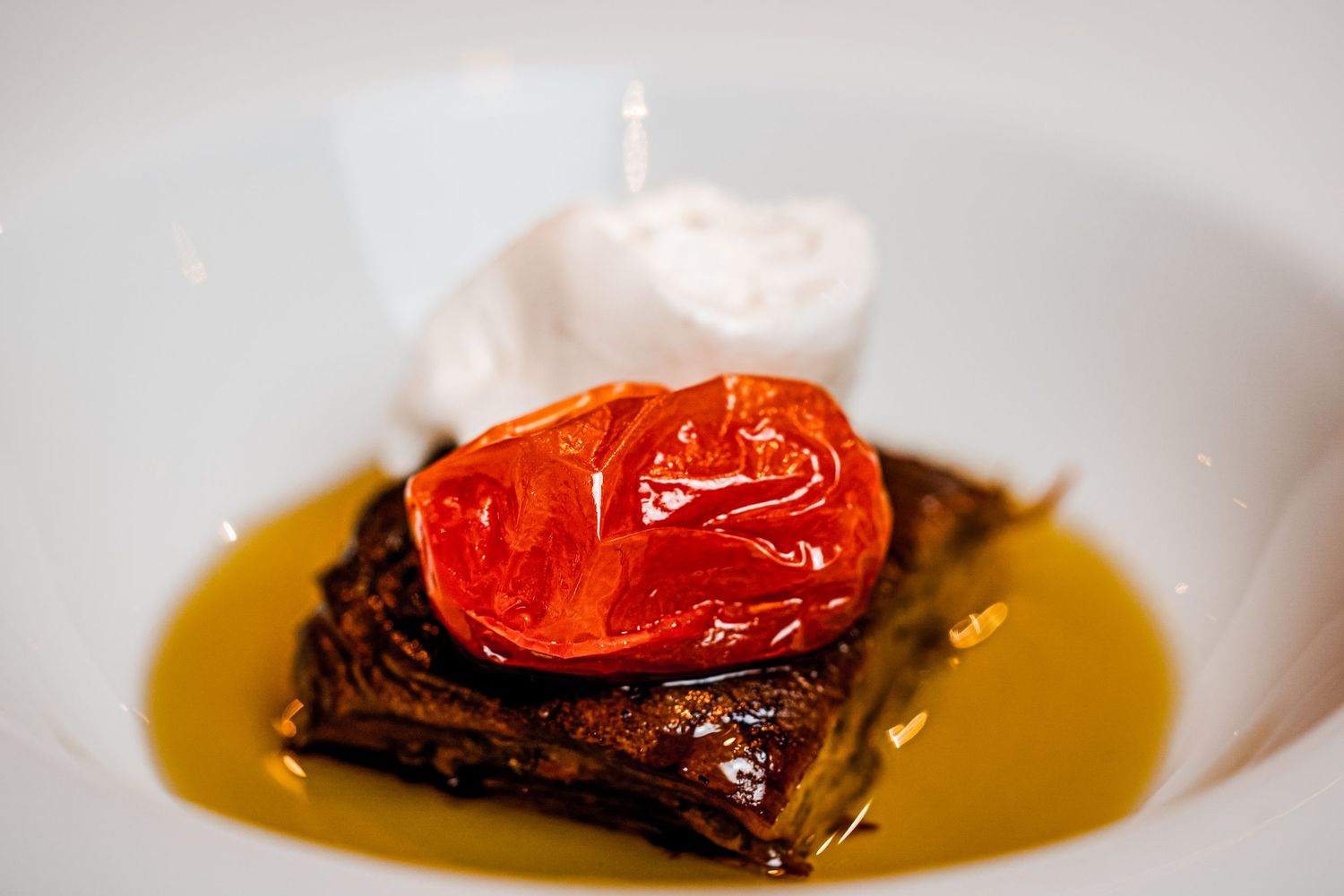 Gourmet dish with roasted tomato on braised meat, olive oil sauce, and whipped cream on white plate.