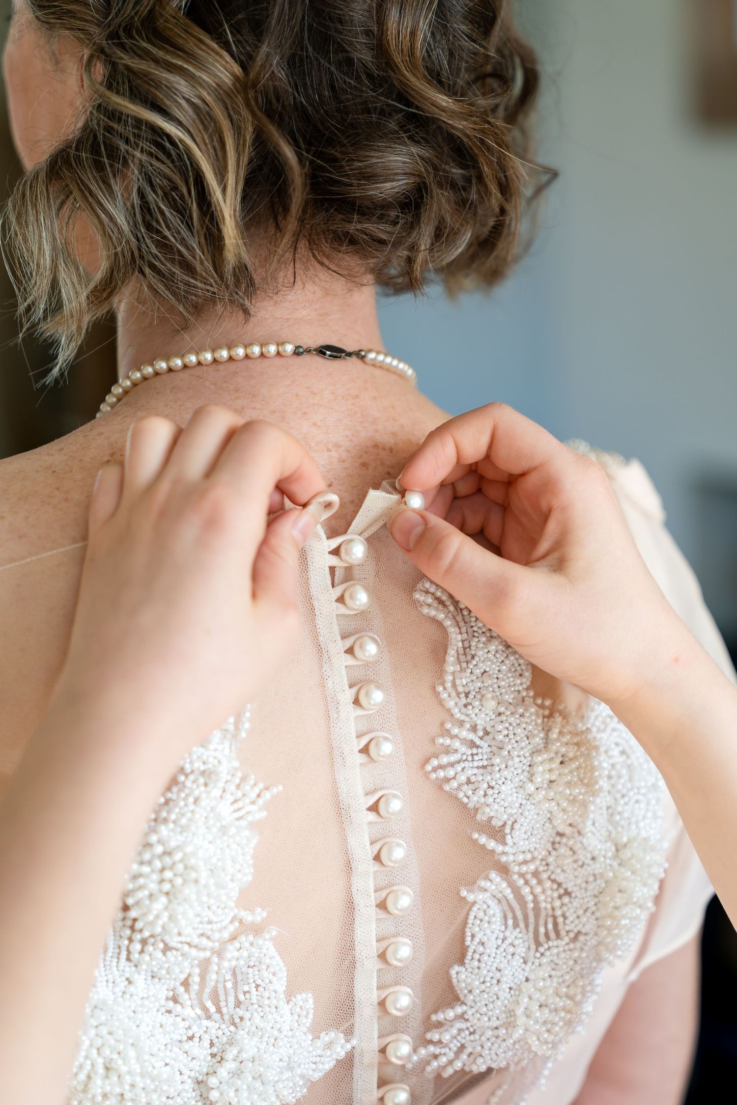 Hands fastening pearl buttons on back of bridal lace gown, bride wearing pearl necklace with elegant updo hairstyle.