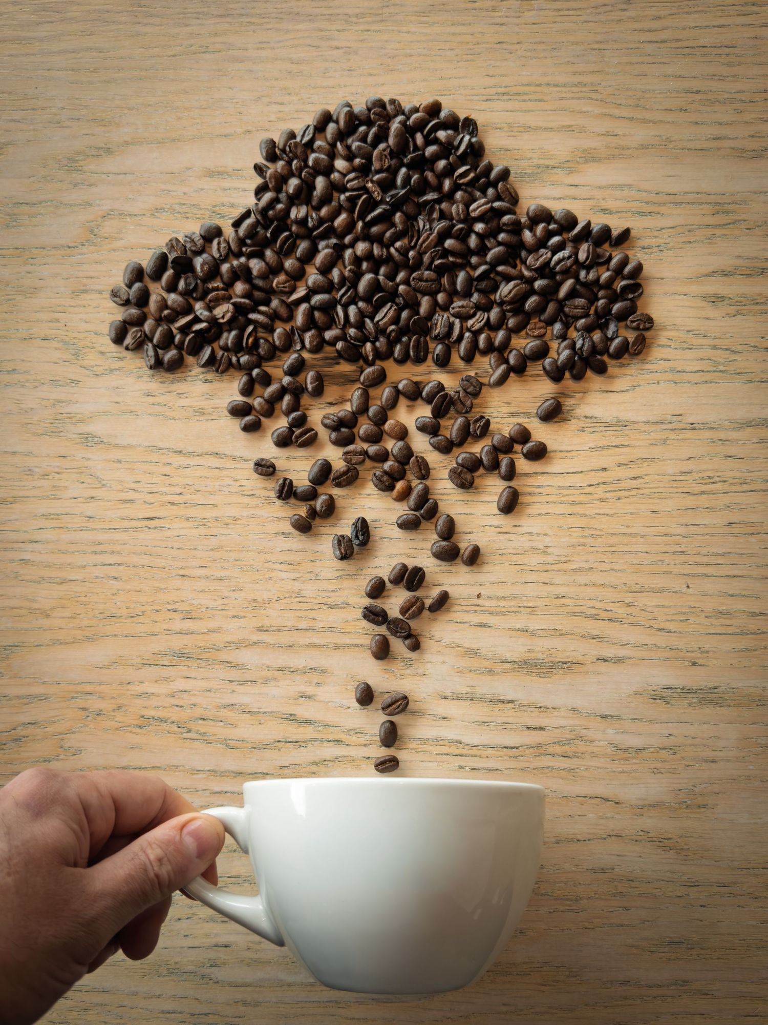 Hand holding white coffee cup with coffee beans arranged as a rain cloud on wooden surface.