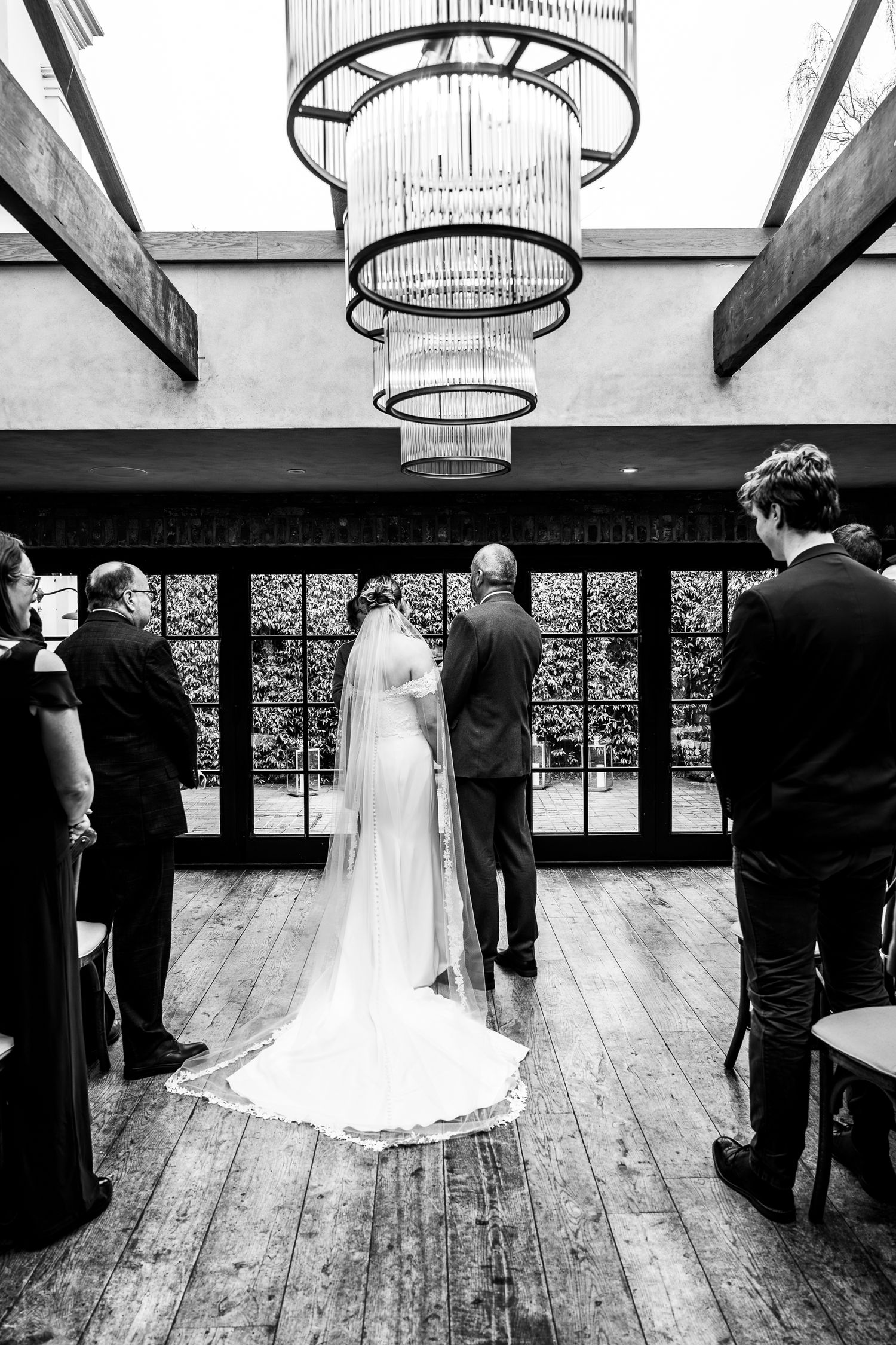 Black and white photo of a bride in a long veil walking down the aisle with her father at an elegant indoor wedding.