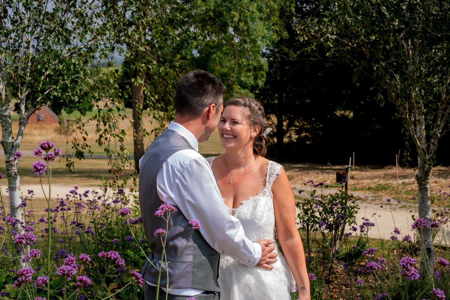 A smiling bride in a white dress embraces her groom in a garden with purple flowers on a sunny wedding day.
