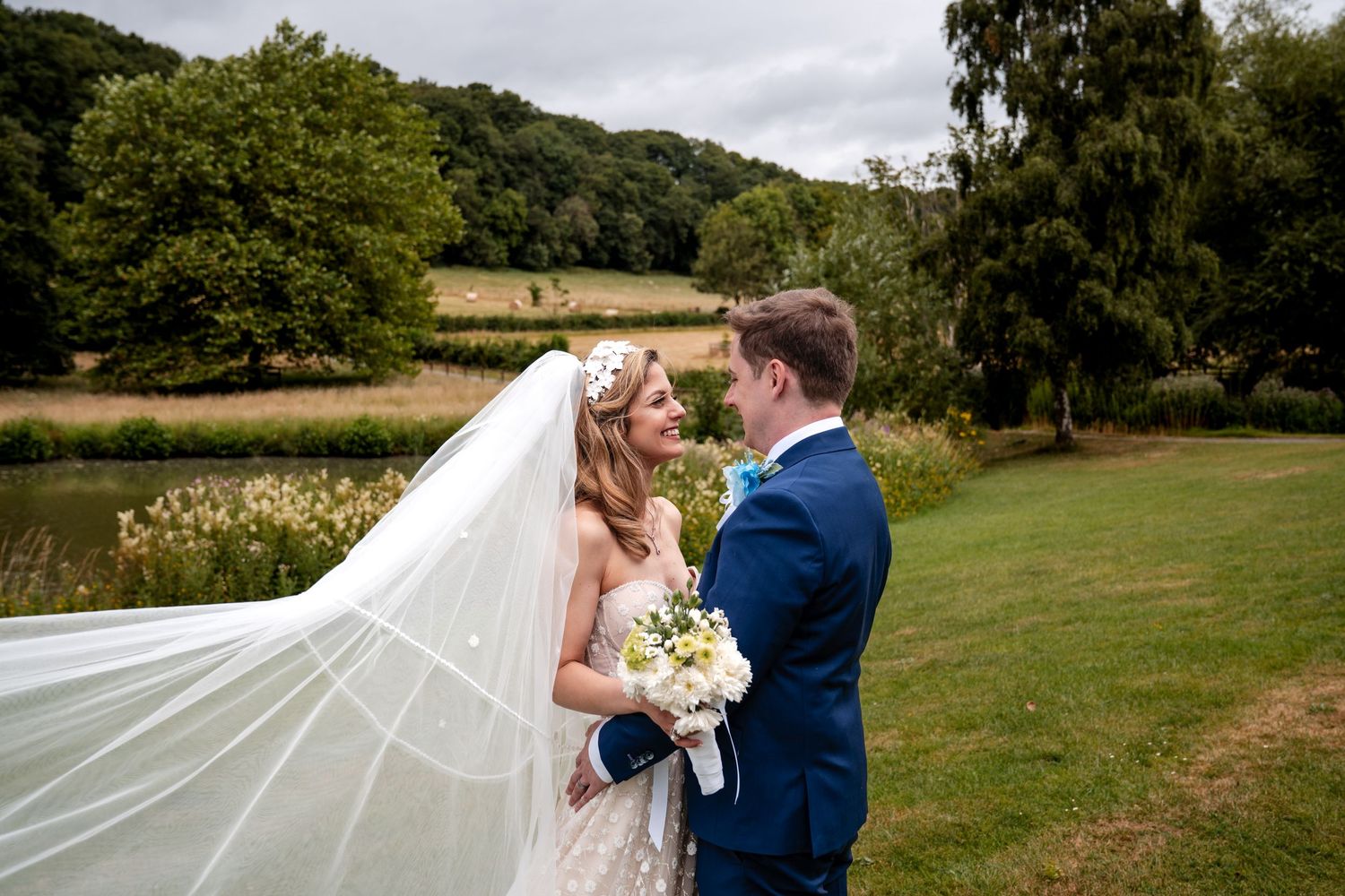 Bride and groom share a romantic moment outdoors, her veil flowing in the wind with a scenic pond and trees behind them.