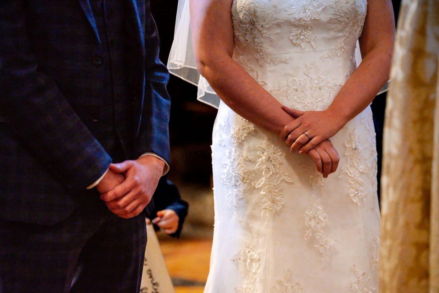 Close-up of bride in lace wedding dress and groom in navy suit holding hands during wedding ceremony.