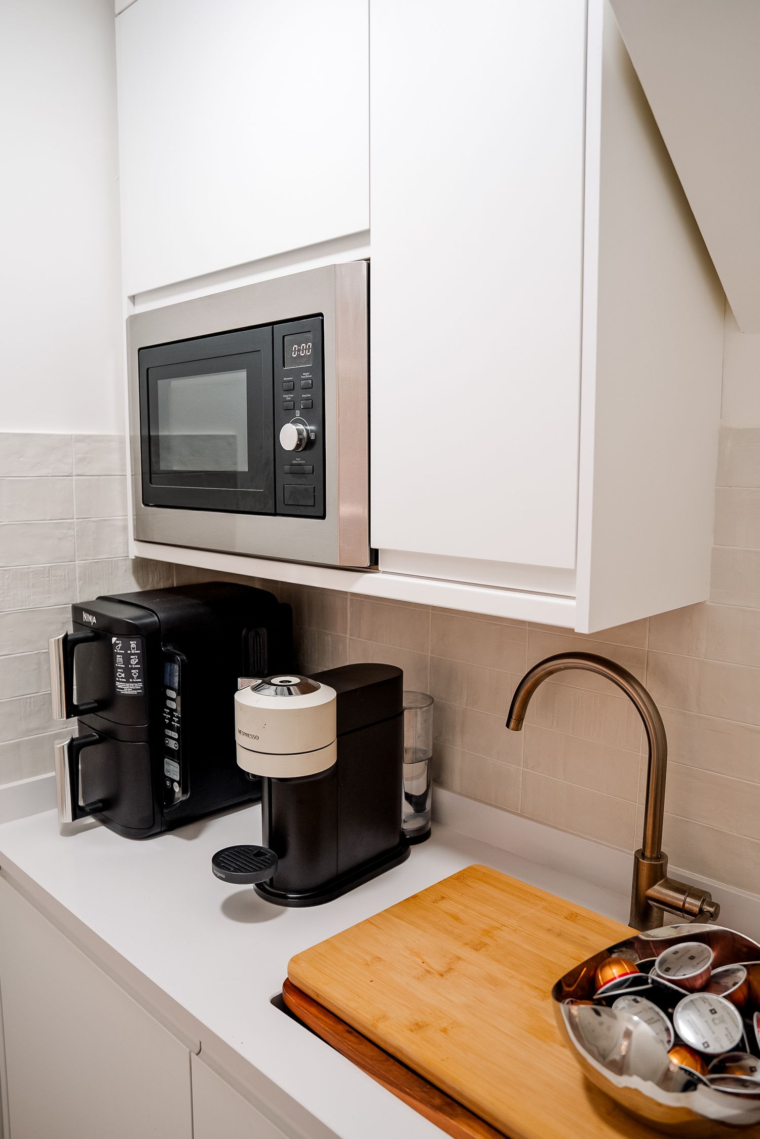 Modern white kitchen counter with built-in microwave, coffee makers, gold faucet, and wooden cutting board.