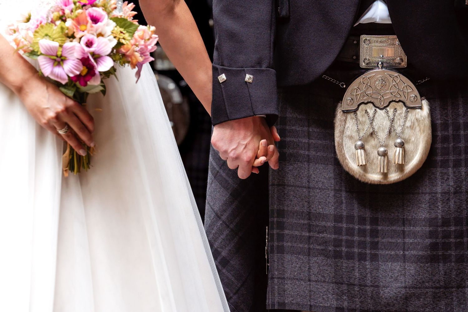 Bride and groom holding hands, wearing wedding dress and Scottish tartan kilt with sporran, bride holding pink floral bouquet.