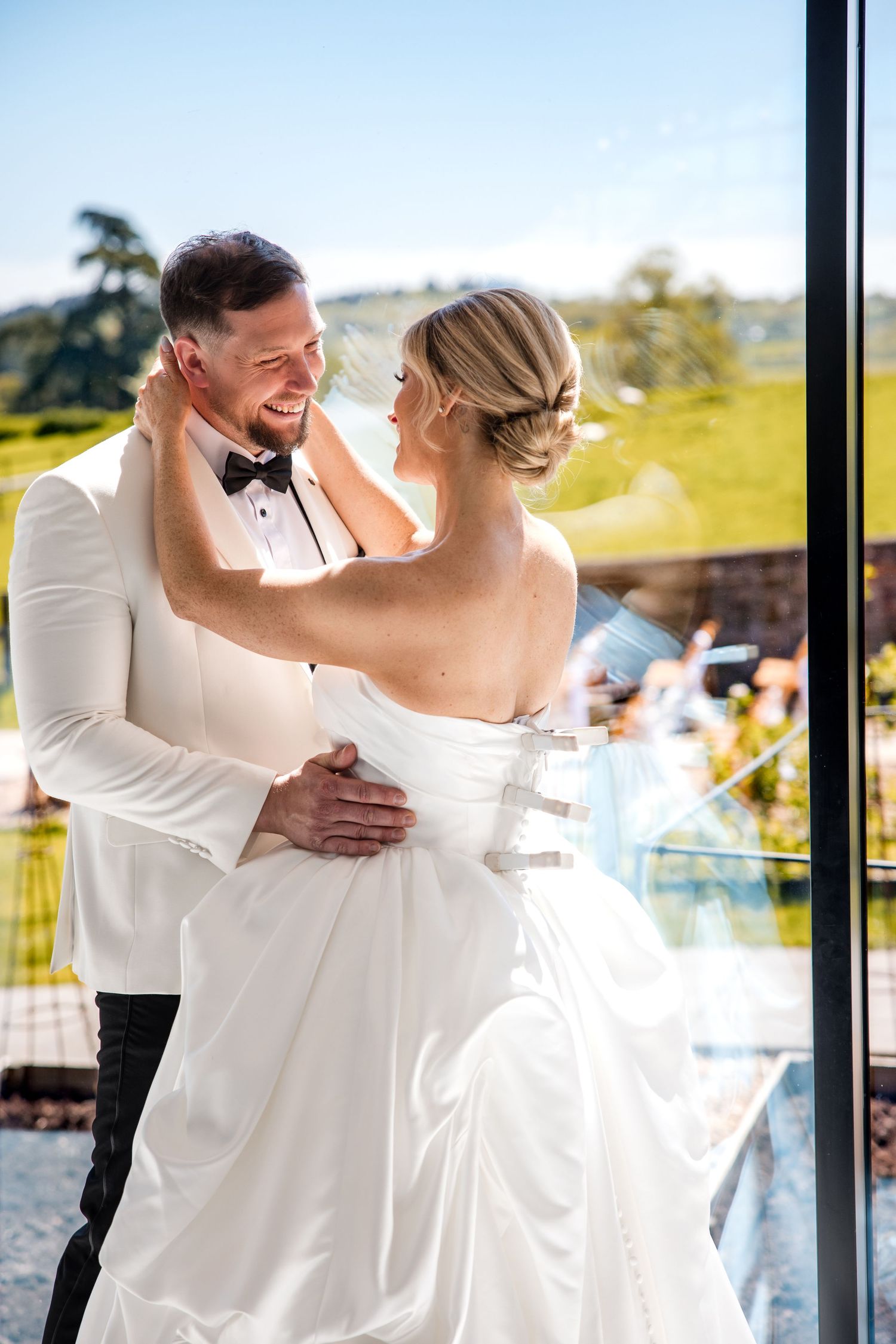A smiling bride and groom in white wedding attire share a romantic embrace near a glass window with green hills behind.