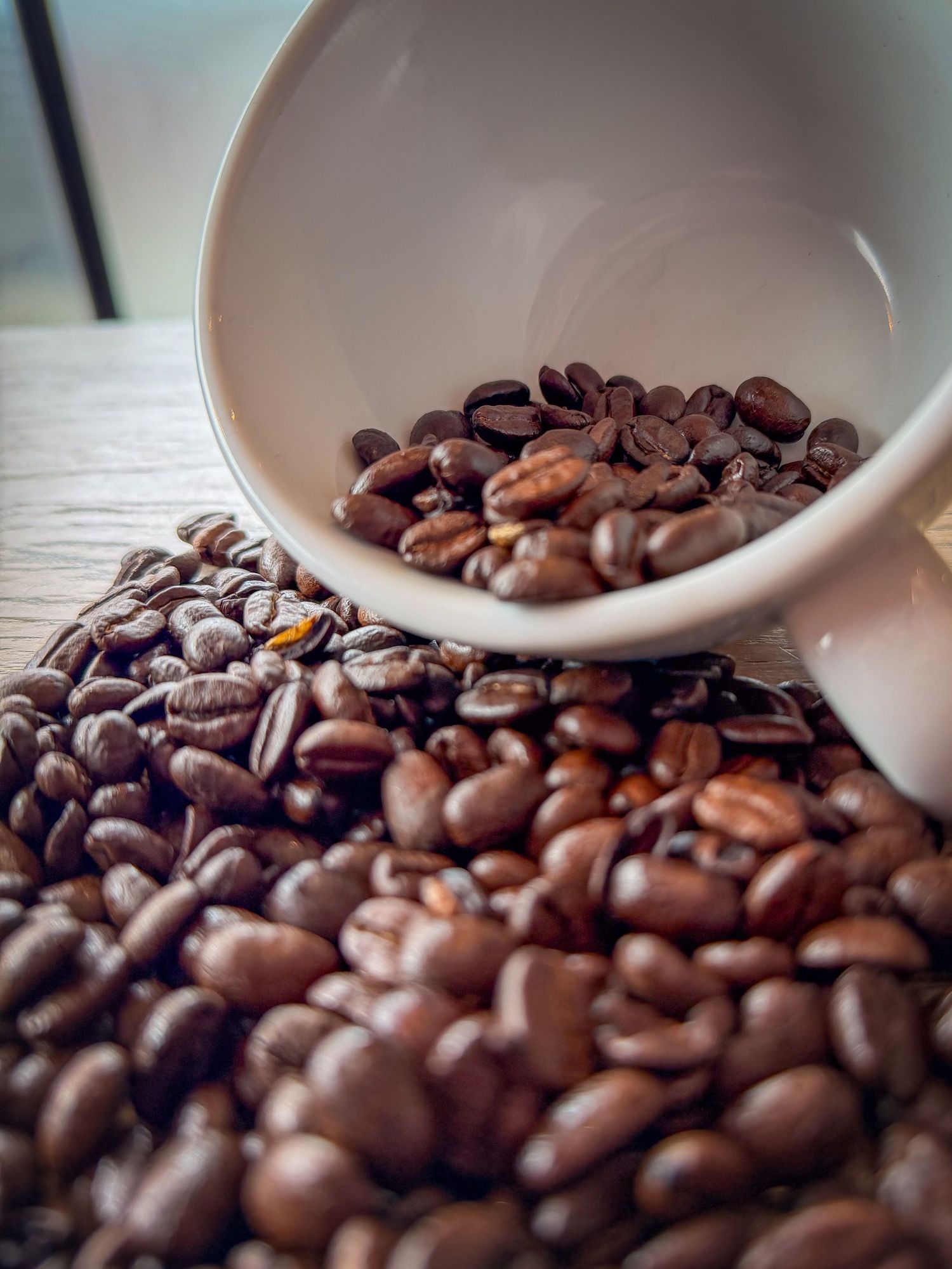 A white ceramic cup tipping over and spilling roasted coffee beans onto a wooden surface.
