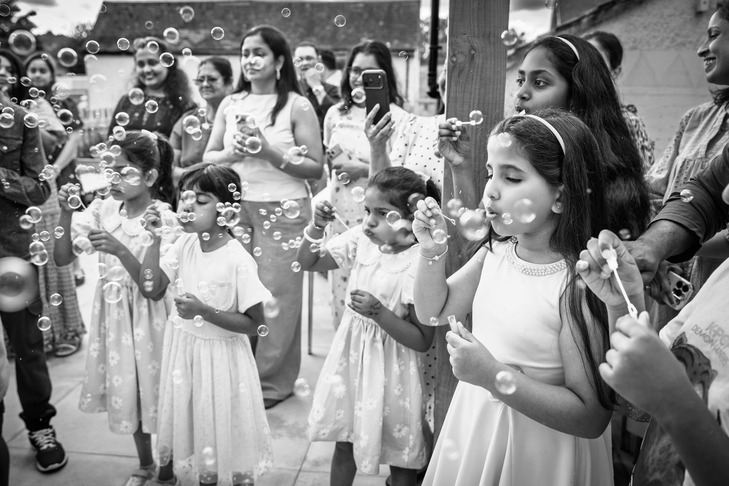 Black and white photo of children in white dresses blowing bubbles at a festive outdoor celebration.