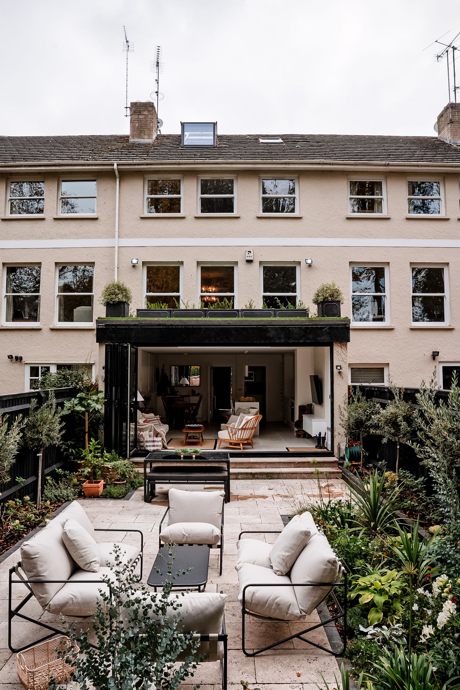 Elegant townhouse garden patio with cream outdoor sofas, lush greenery, and open glass doors leading to a stylish interior.
