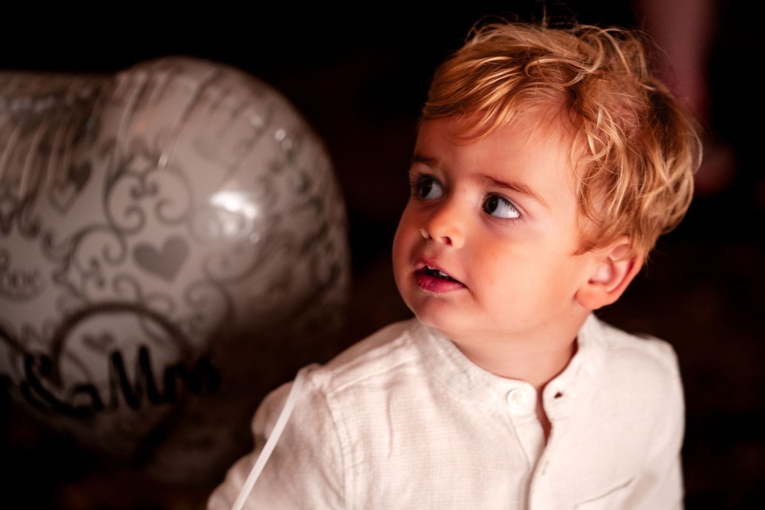 Young blonde toddler boy in white shirt gazes upward curiously beside a decorative balloon at a celebration.