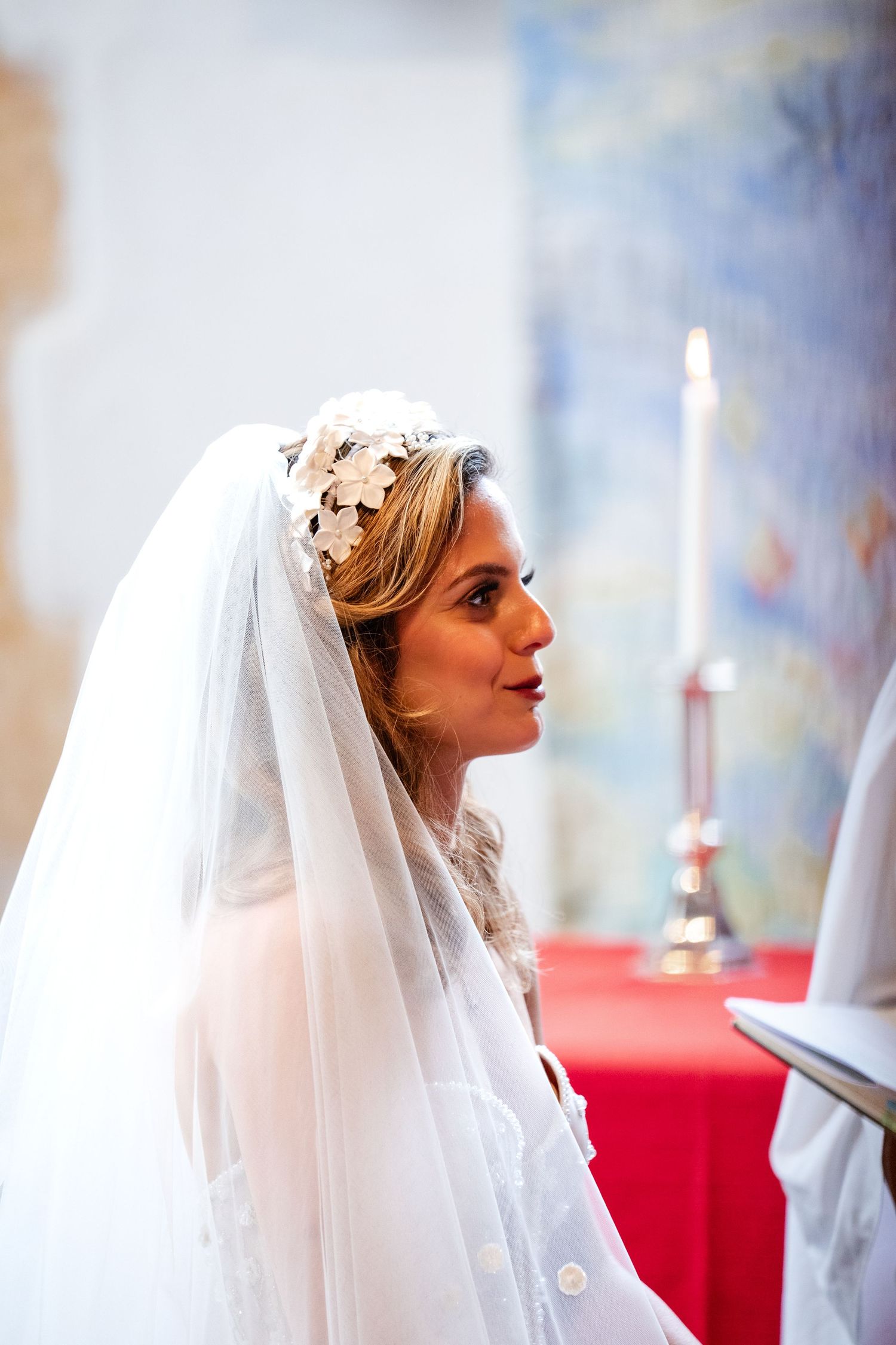 Bride in white veil and floral headpiece stands at church altar during wedding ceremony with candles in background.