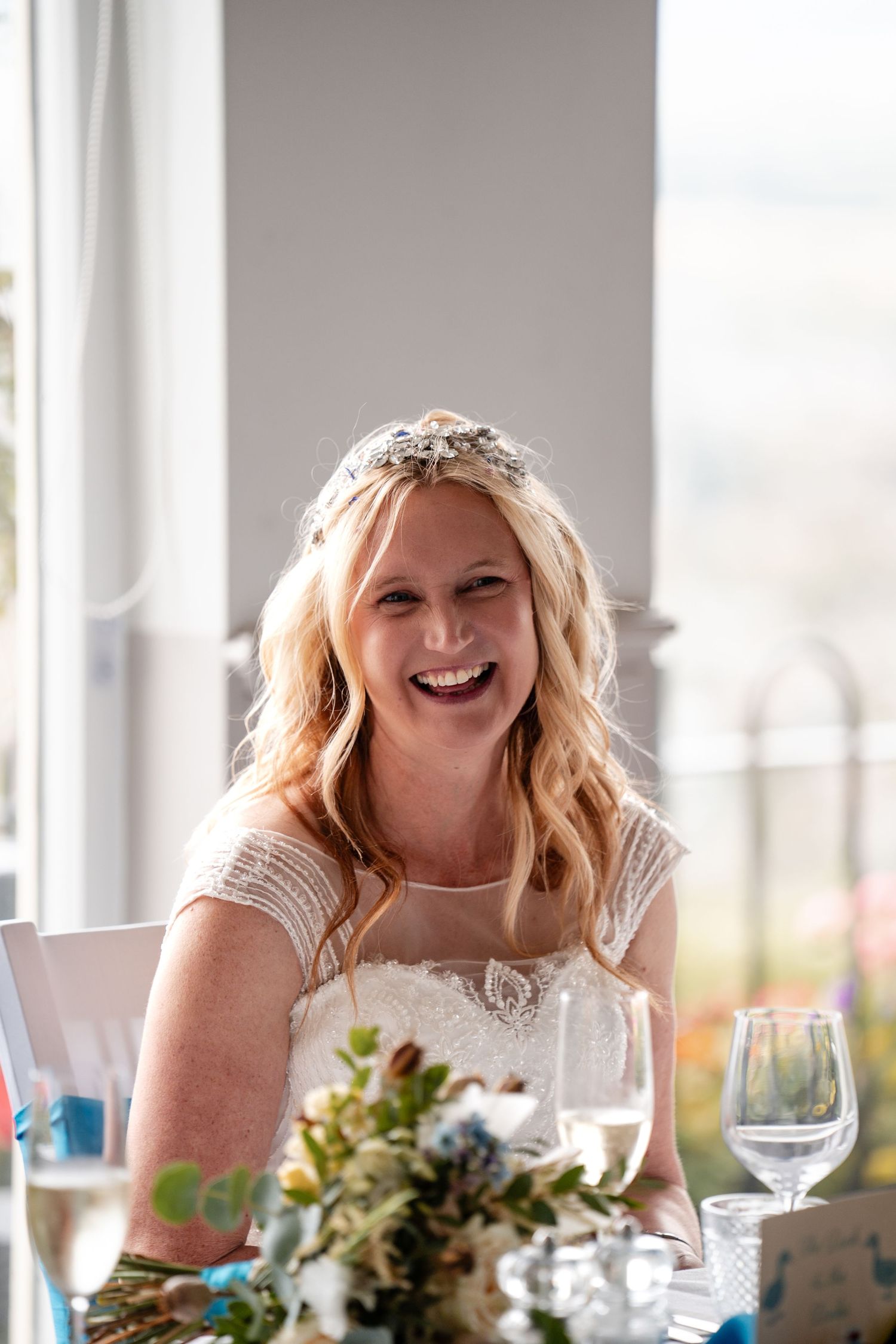 Smiling bride in white lace dress and floral crown laughing joyfully at wedding reception table with flowers.