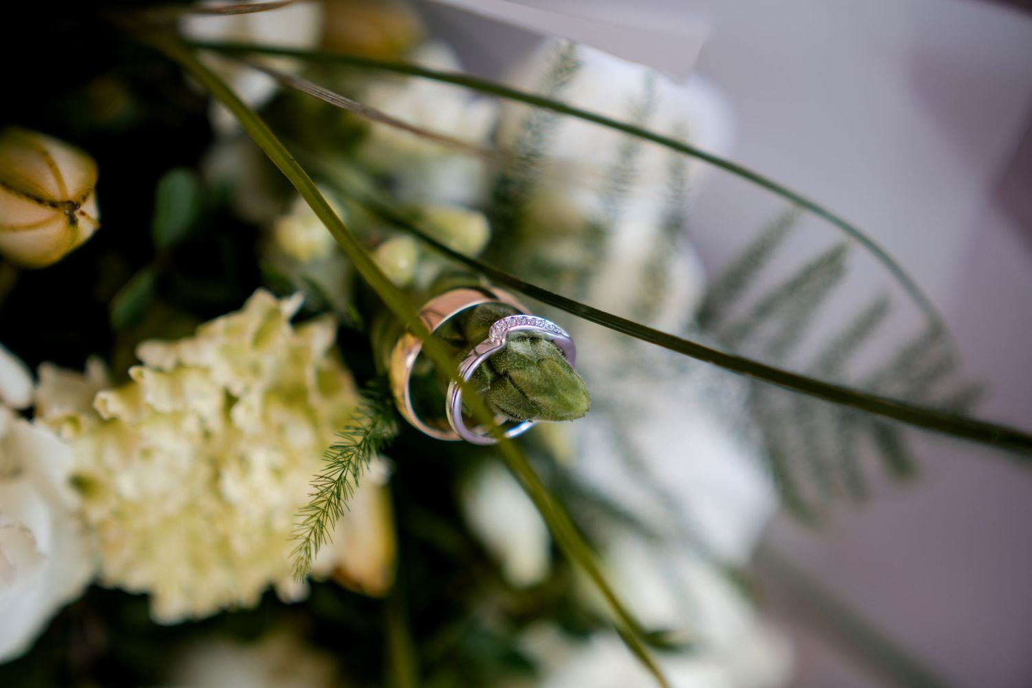 Two wedding rings resting on a green stem surrounded by white flowers and lush greenery in a bridal bouquet.