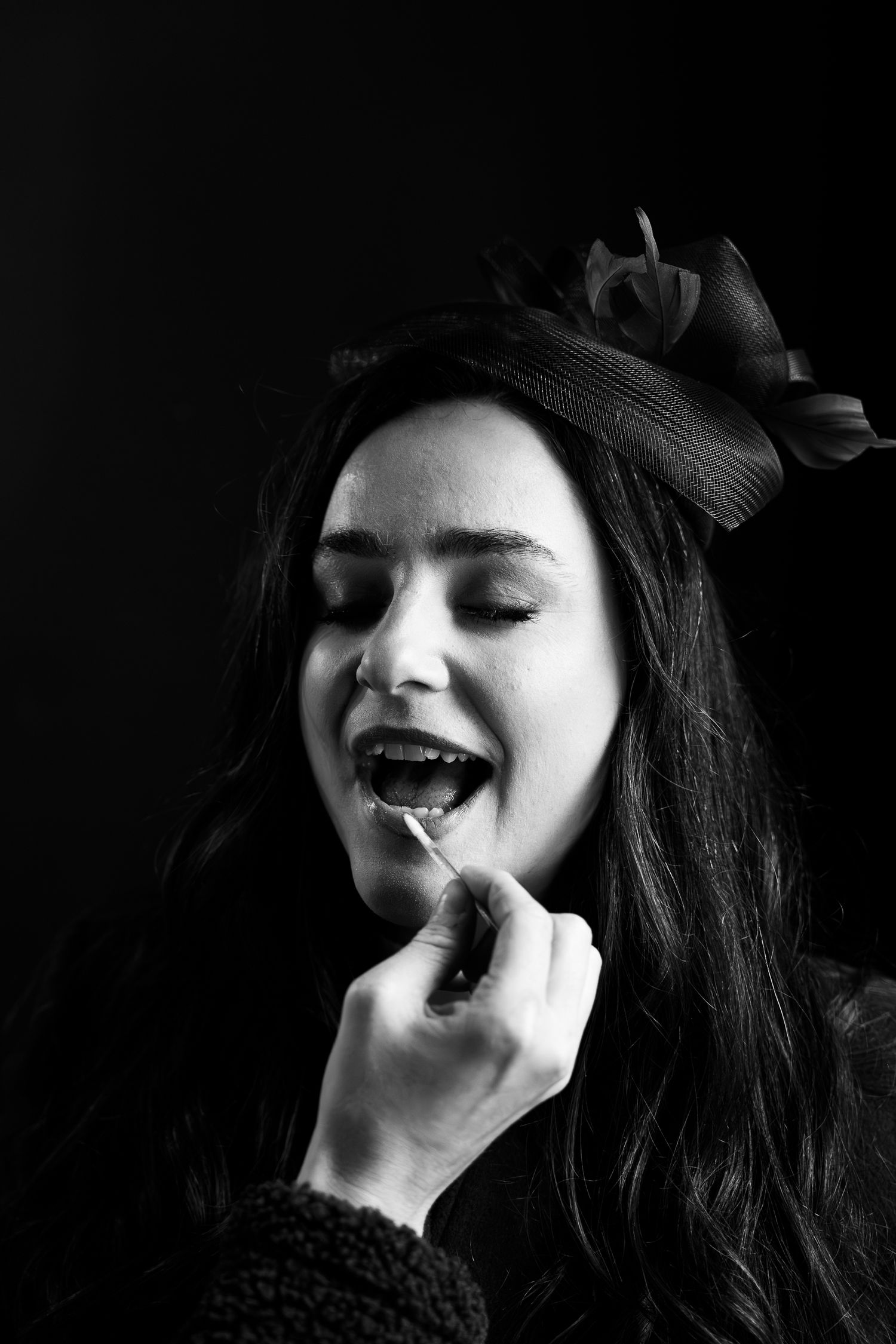 Black and white portrait of a woman with hair bow having lipstick applied with a brush, eyes closed.