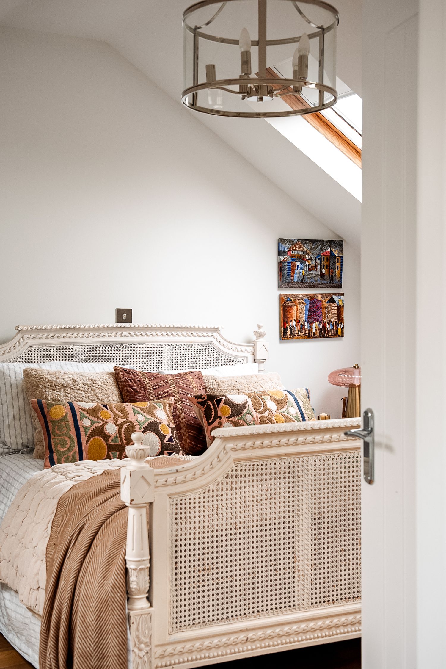 Attic bedroom with cane bed frame, earth-tone pillows, skylight, lantern chandelier, and colorful wall art.
