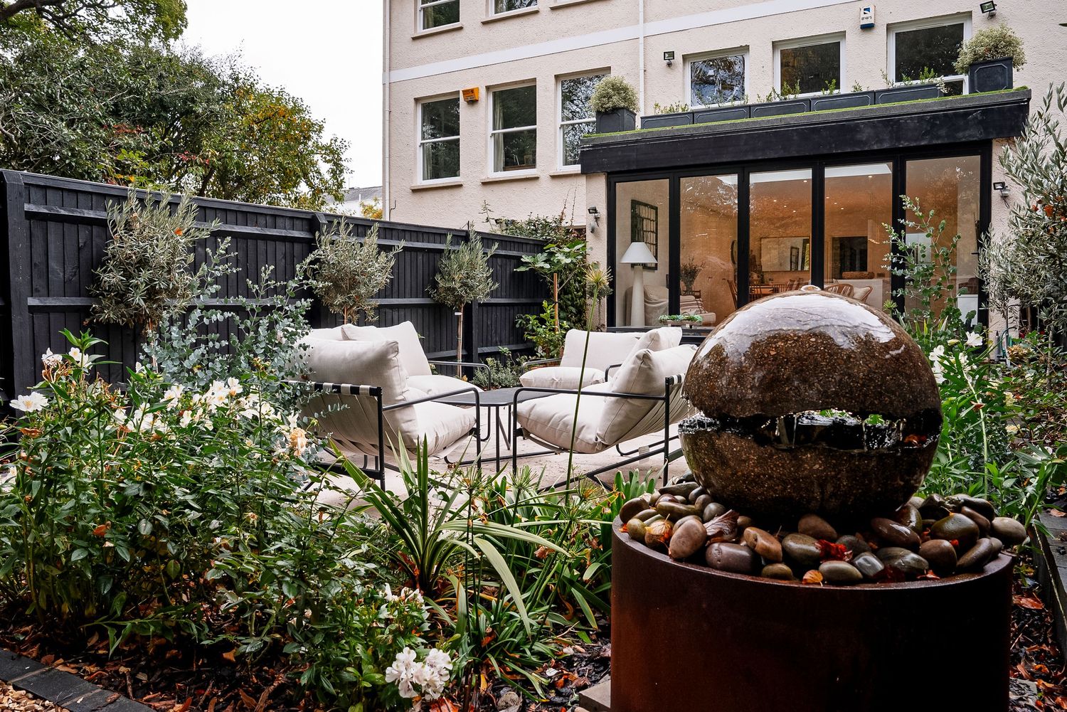 Lush garden patio with stone sphere water fountain, white outdoor seating, and modern glass extension in background.