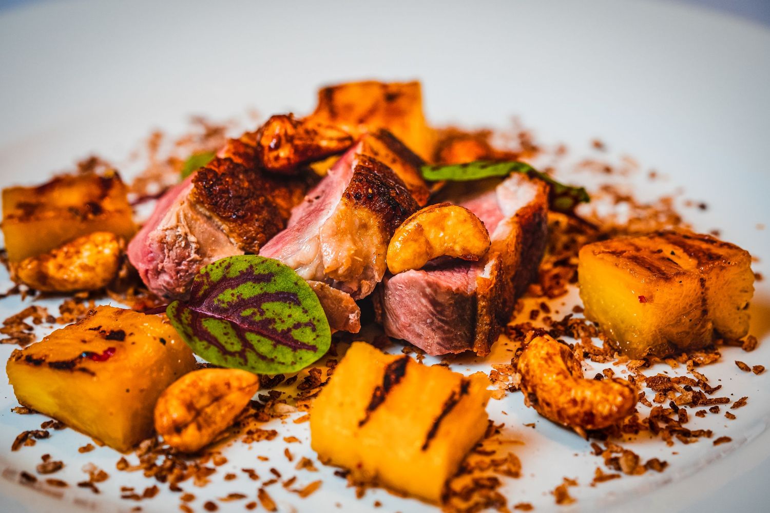 Gourmet plated dish with seared duck breast, caramelized pineapple chunks, roasted nuts, and fresh herbs on white plate.