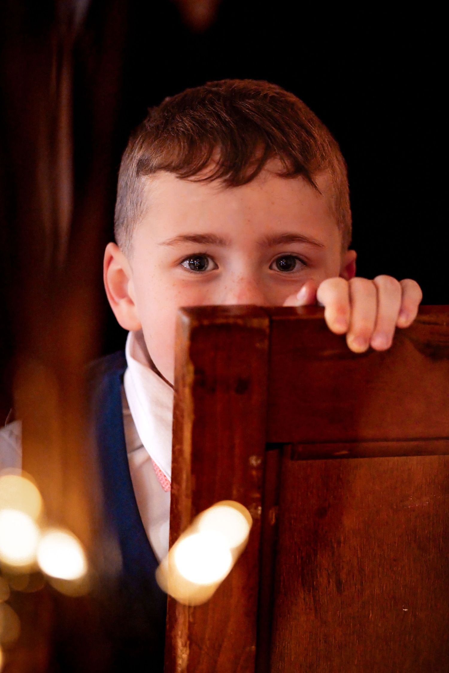 Young boy peeks over a wooden church pew with curious eyes, surrounded by soft candlelight in a dimly lit church.