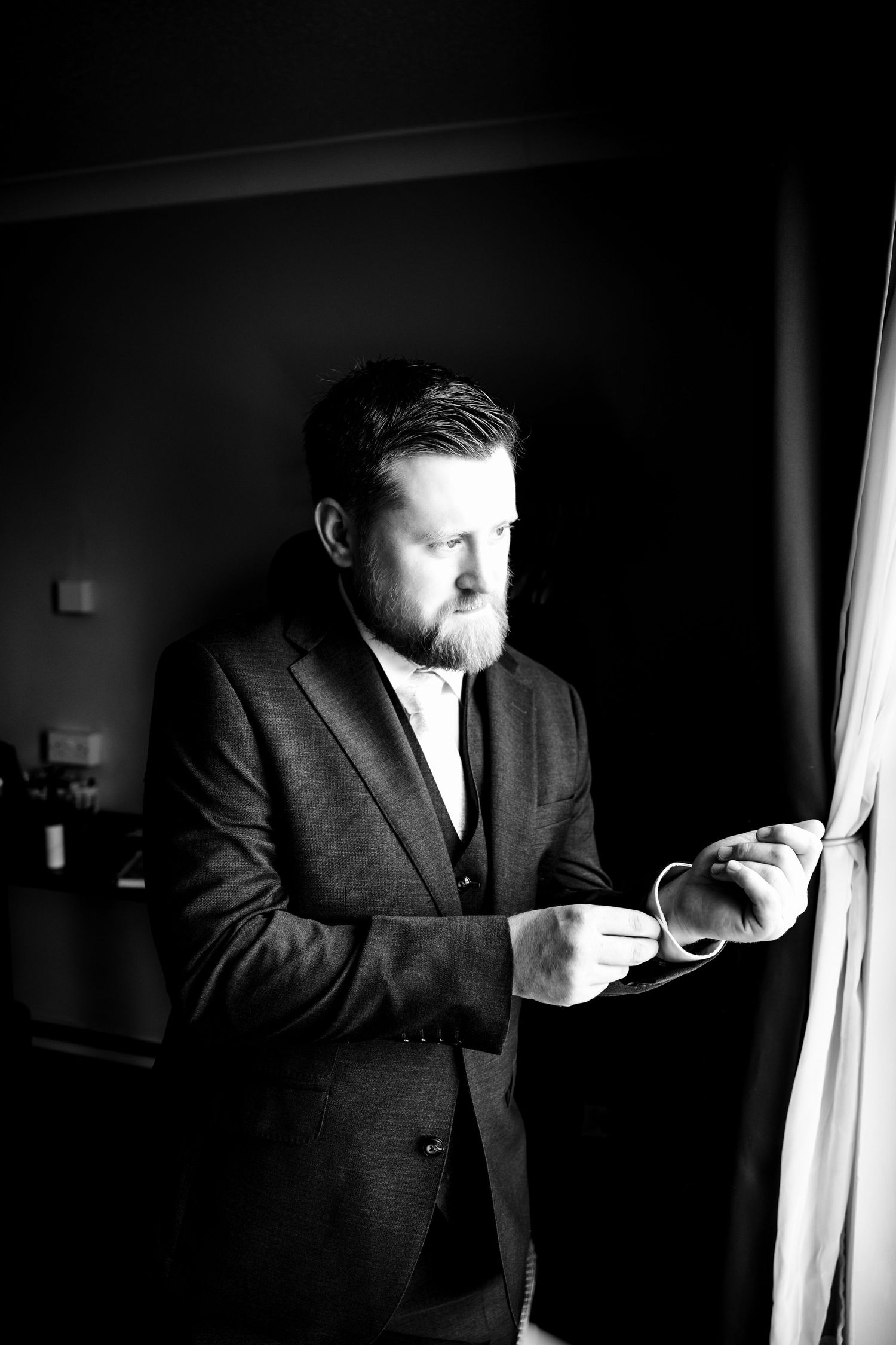 Black and white portrait of a bearded man in a suit adjusting his cufflinks near a window.