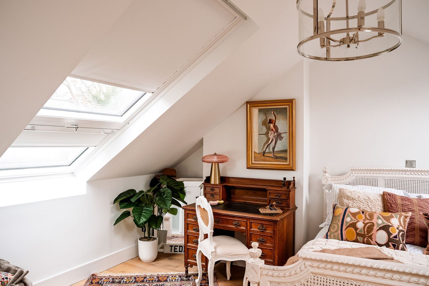Bright attic bedroom with skylights, antique wooden desk, framed artwork, potted plant, and bohemian bedding under a chandelier.