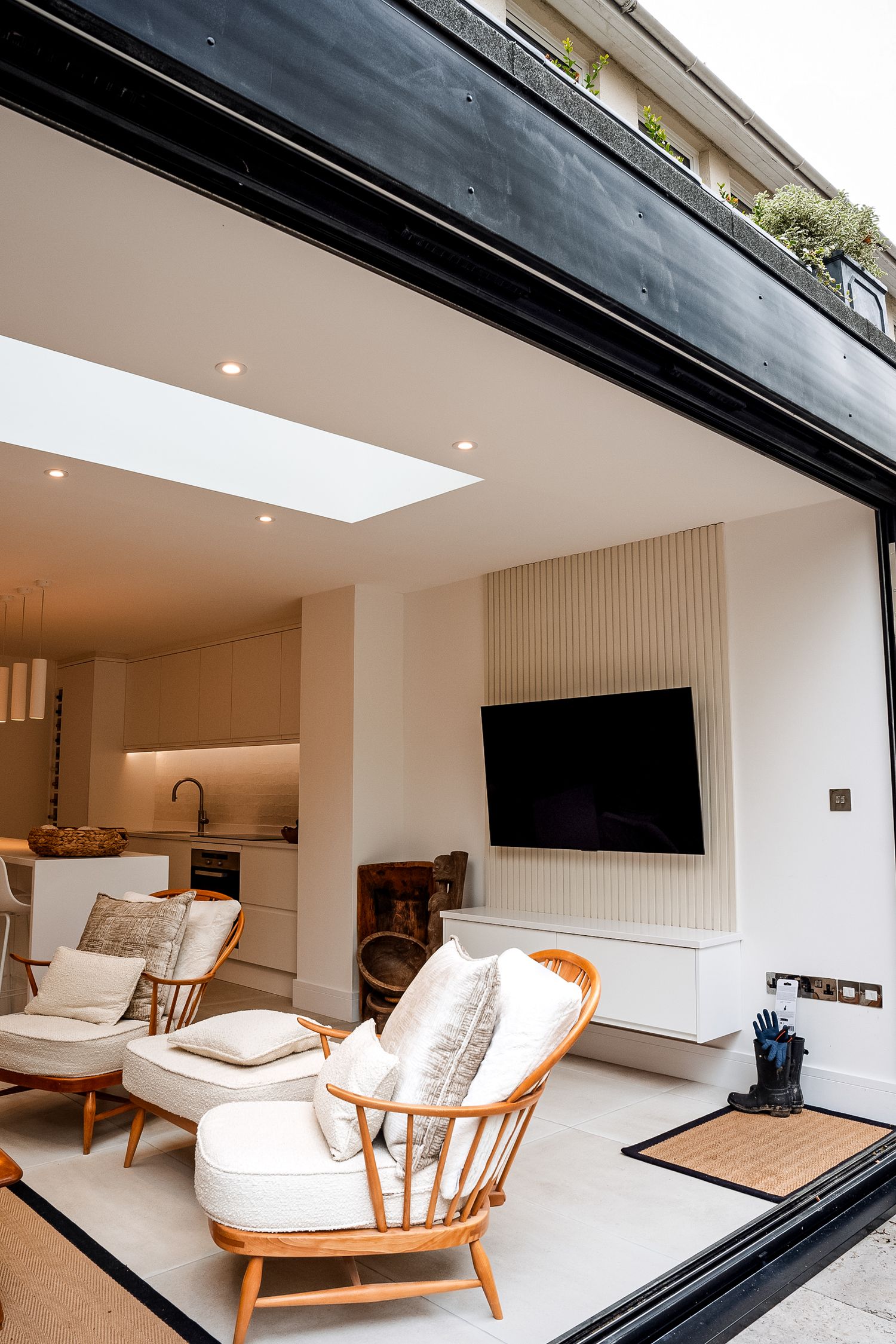 Modern open-plan living room with rattan chairs, wall-mounted TV, skylight, and seamless indoor-outdoor flow via sliding glass doors.