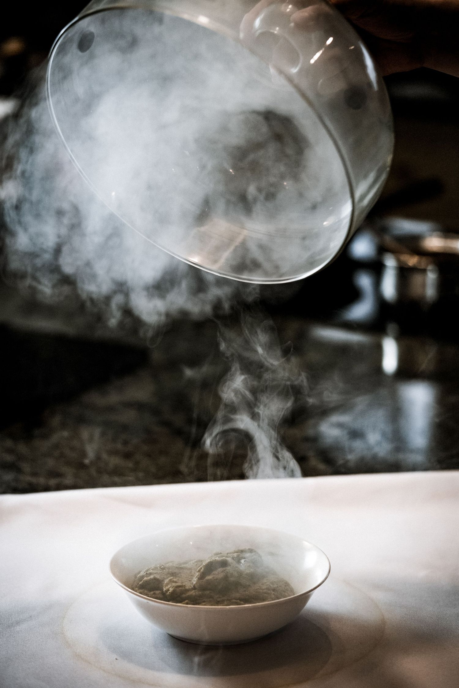 Steam rising dramatically from a bowl of hot soup as a glass lid is lifted in a dark restaurant kitchen.