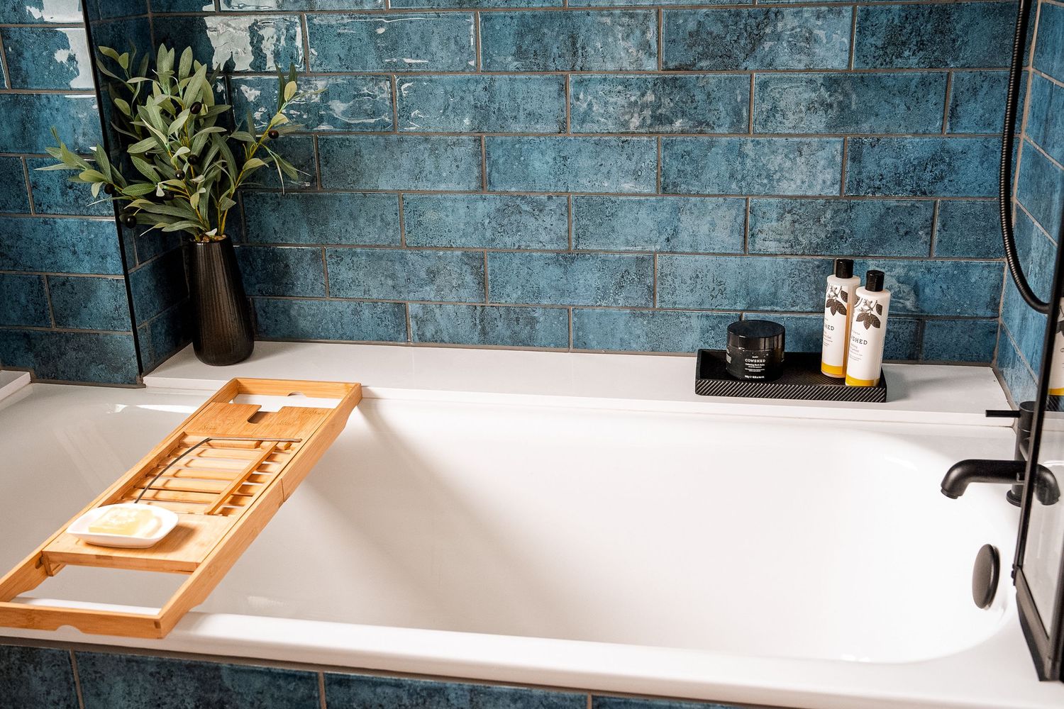 White bathtub with bamboo tray against blue tiled wall, decorated with plants and bath products.