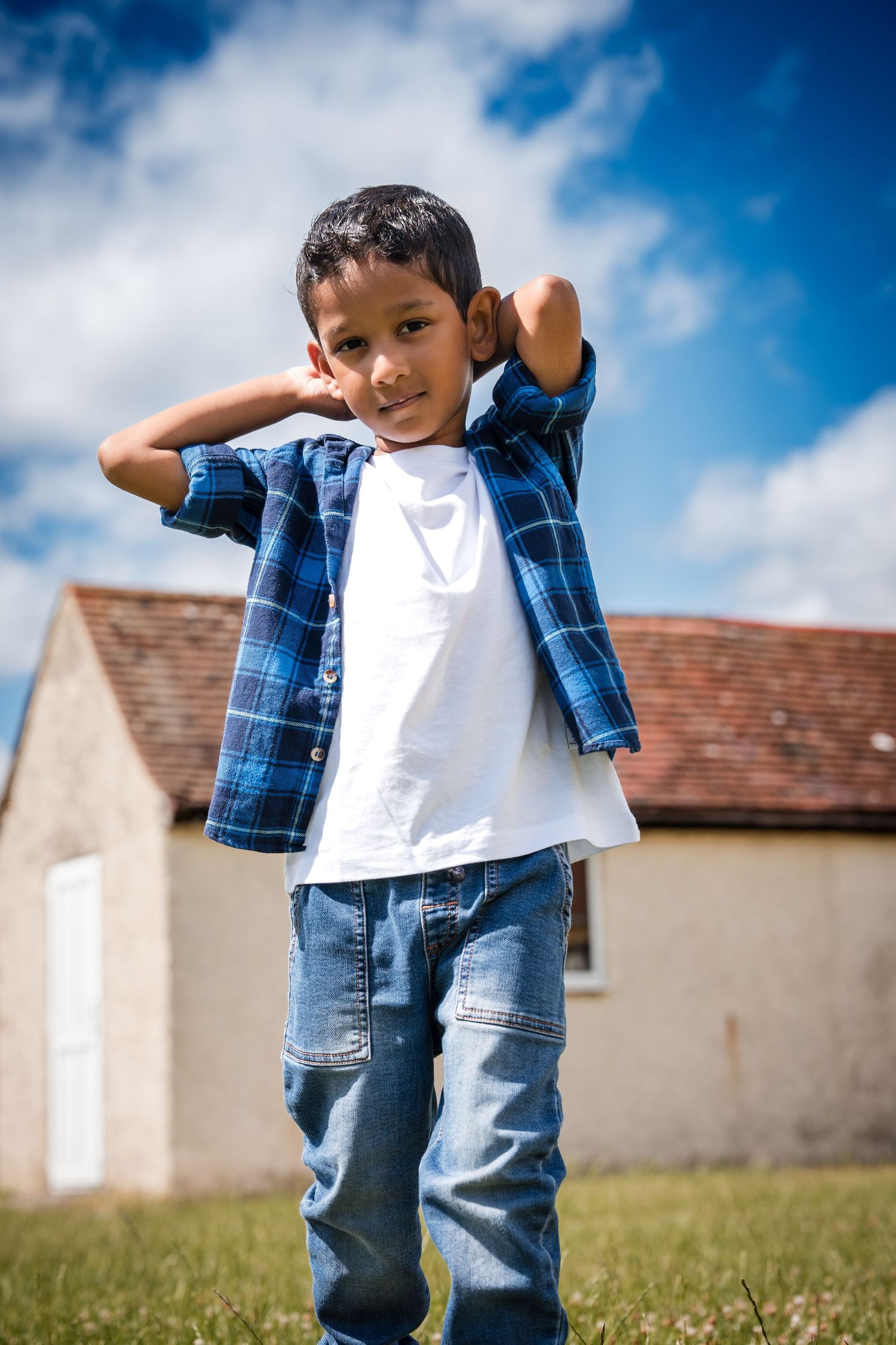 A young boy in a plaid shirt and jeans poses outdoors with hands behind his neck, blue sky and house in background.