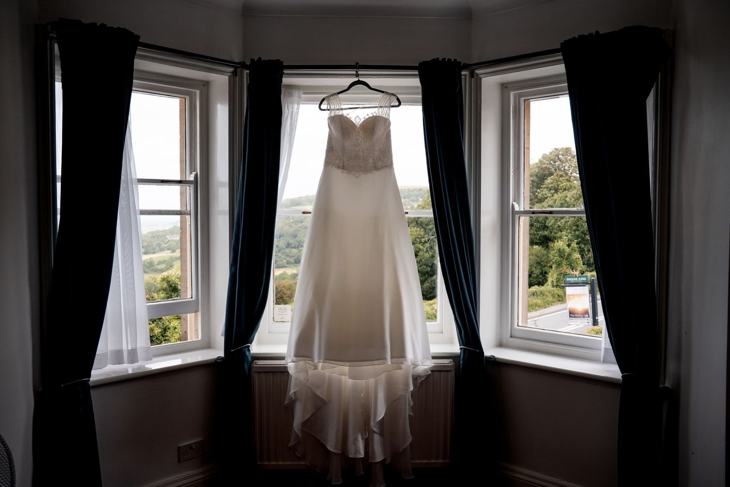 A elegant strapless wedding dress hanging in a bay window with scenic countryside views and dark curtains.
